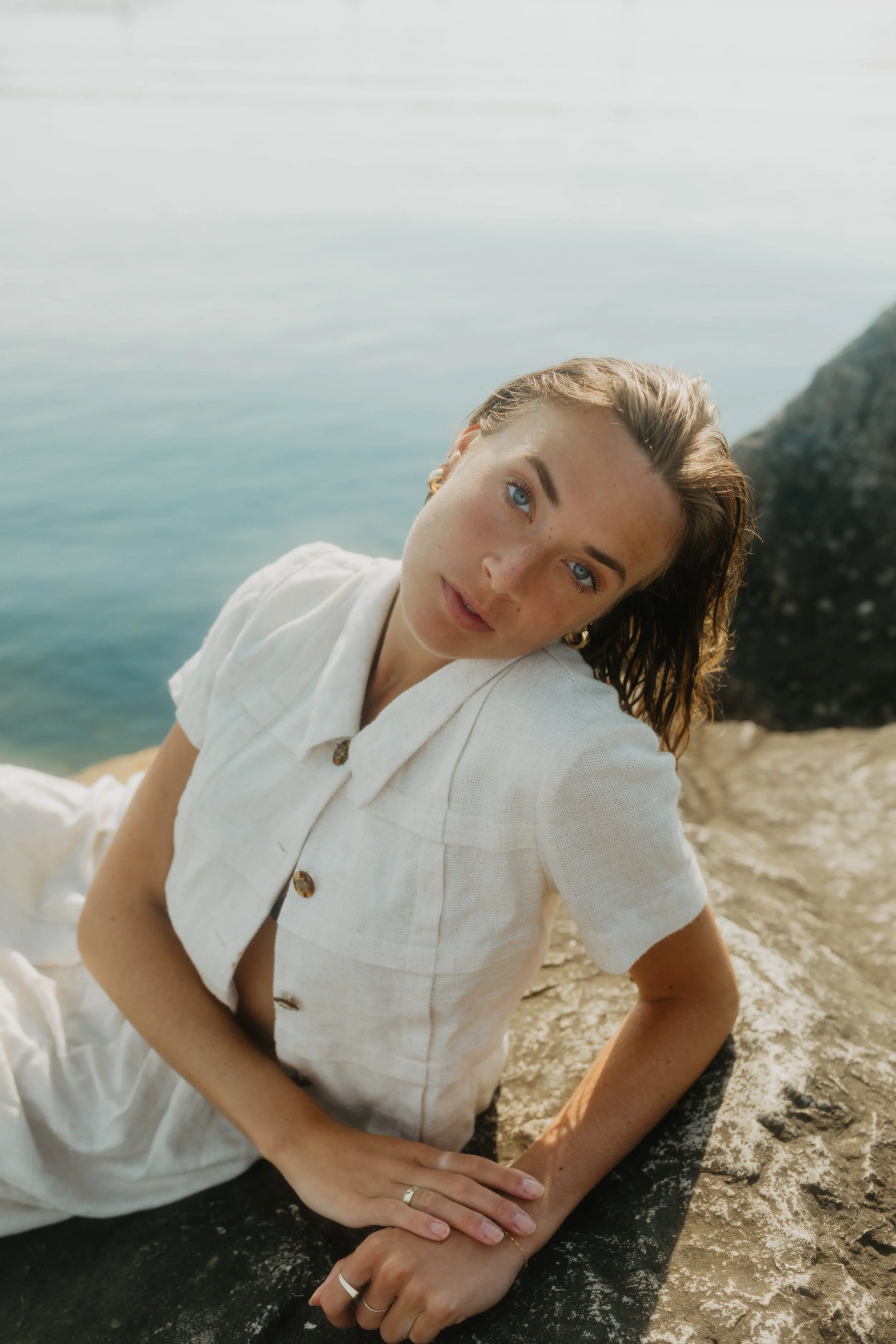 A woman with wet hair and blue eyes lying on a rock near water, wearing a white shirt.