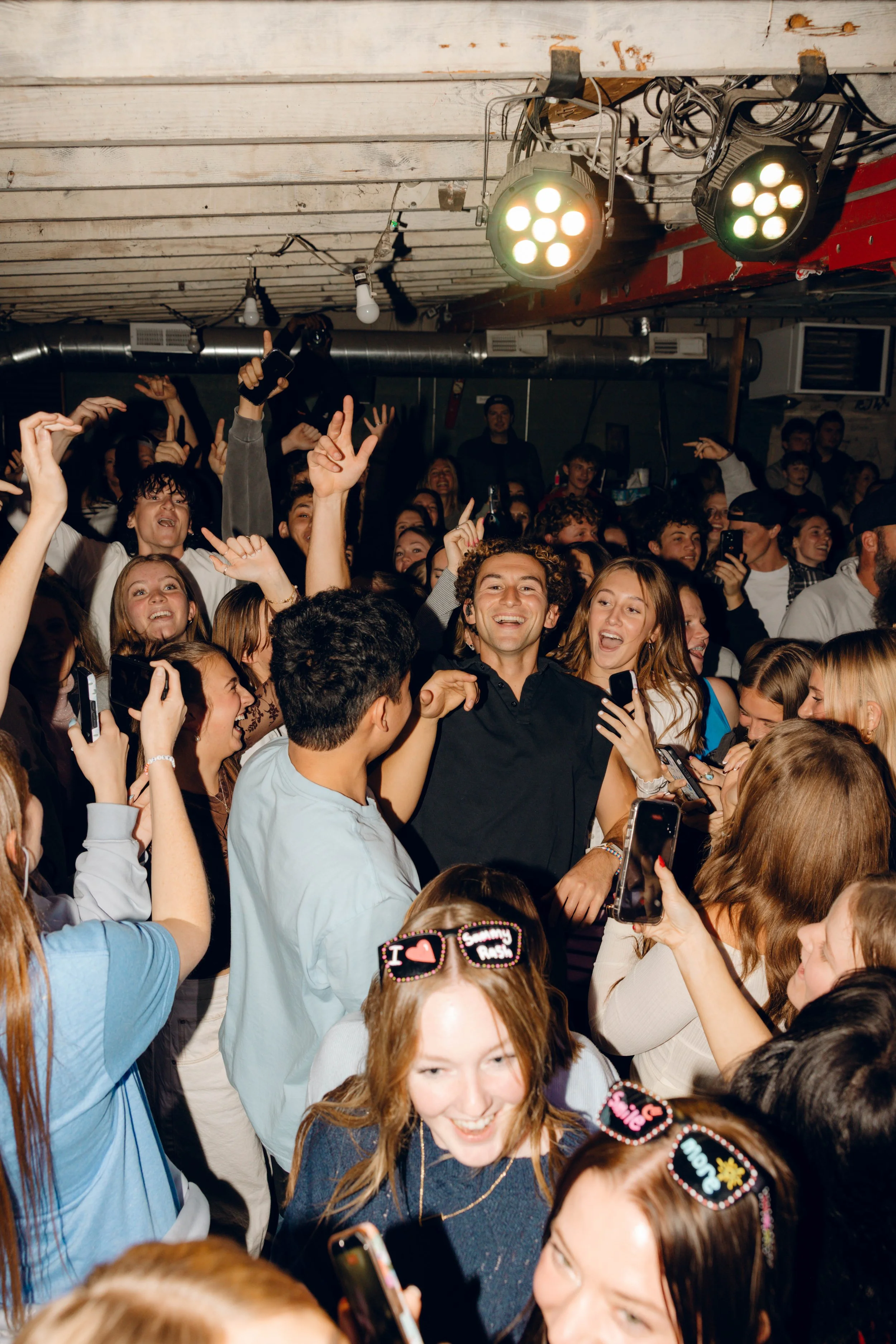 A lively crowd of young people dancing and celebrating in a dimly-lit indoor space, with some holding up phones to take pictures and videos.