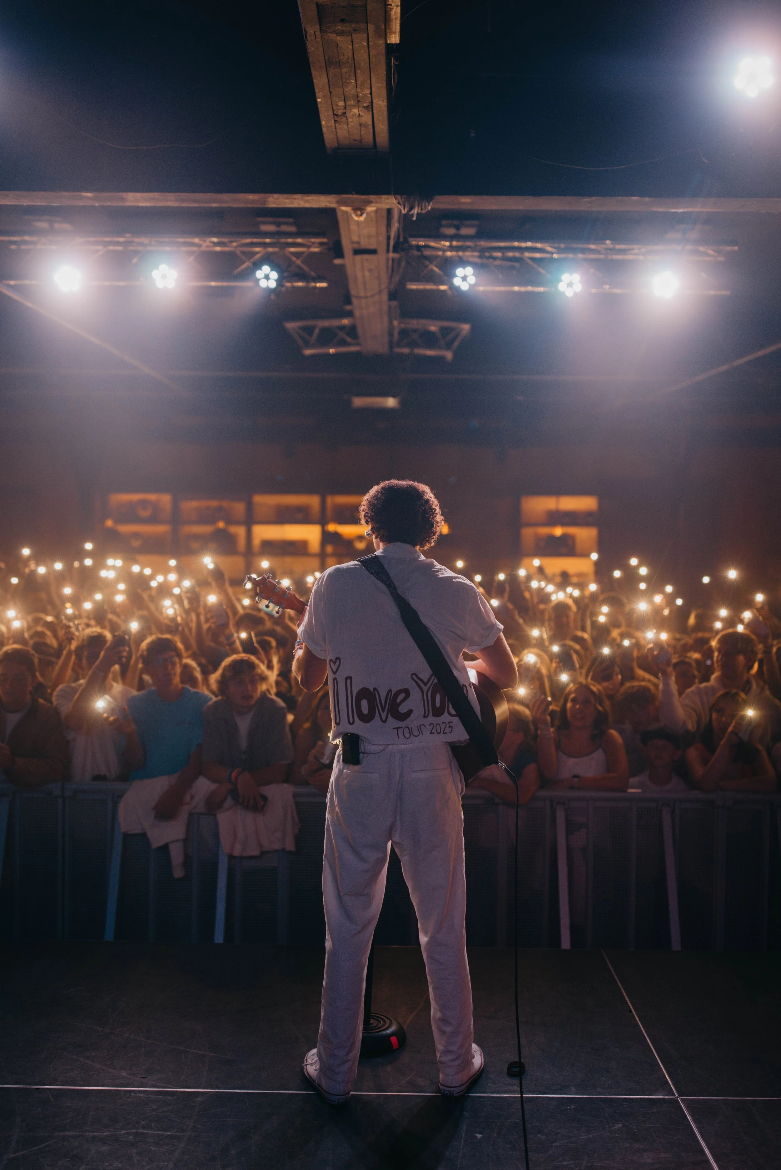 The musician Sammy Rash playing guitar on stage during a concert, facing a large audience holding up phones with lights on, in a dark venue in Salt Lake City.