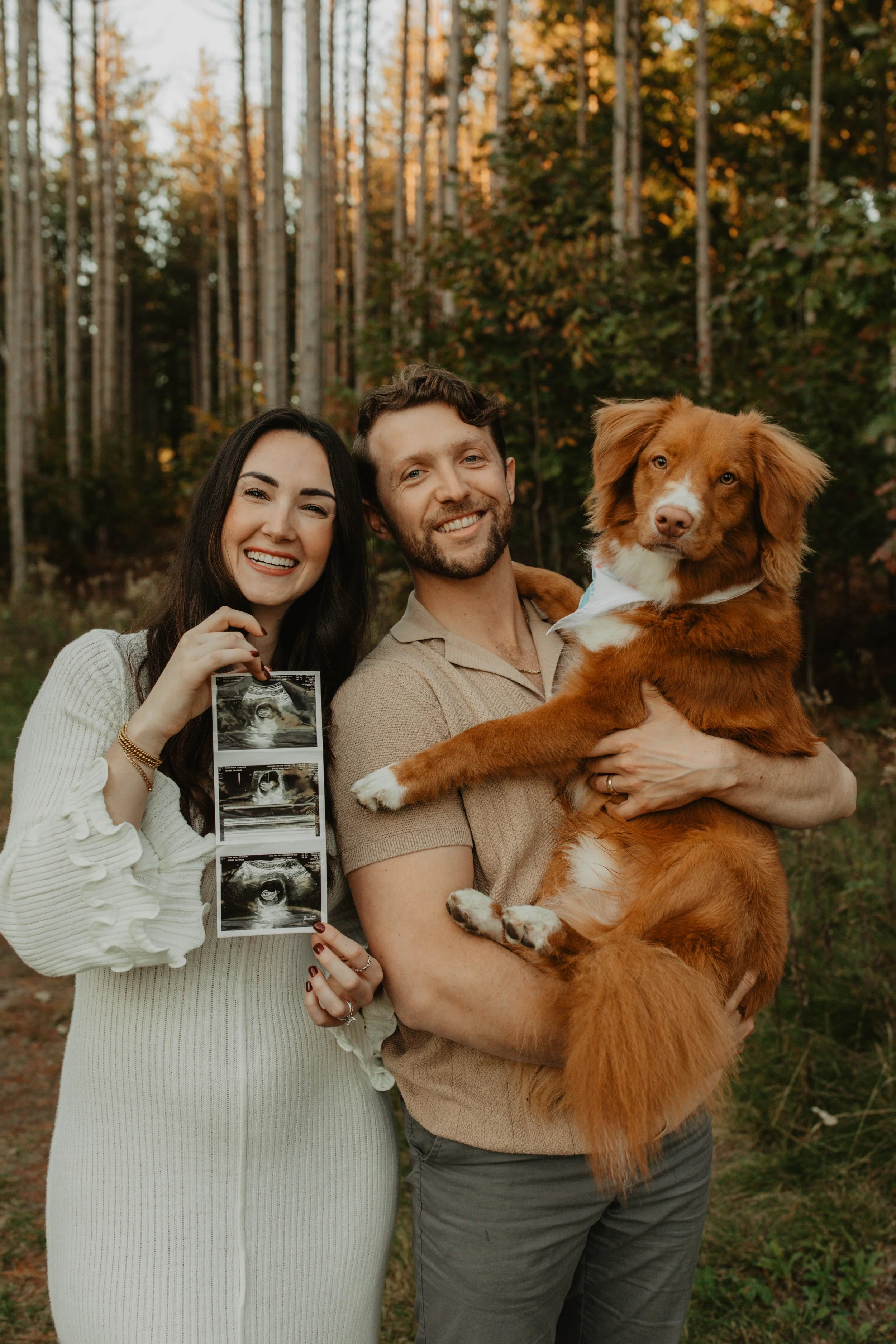 A happy couple holding a large brown dog outdoors in a forest during fall, with the woman holding ultrasound photos.