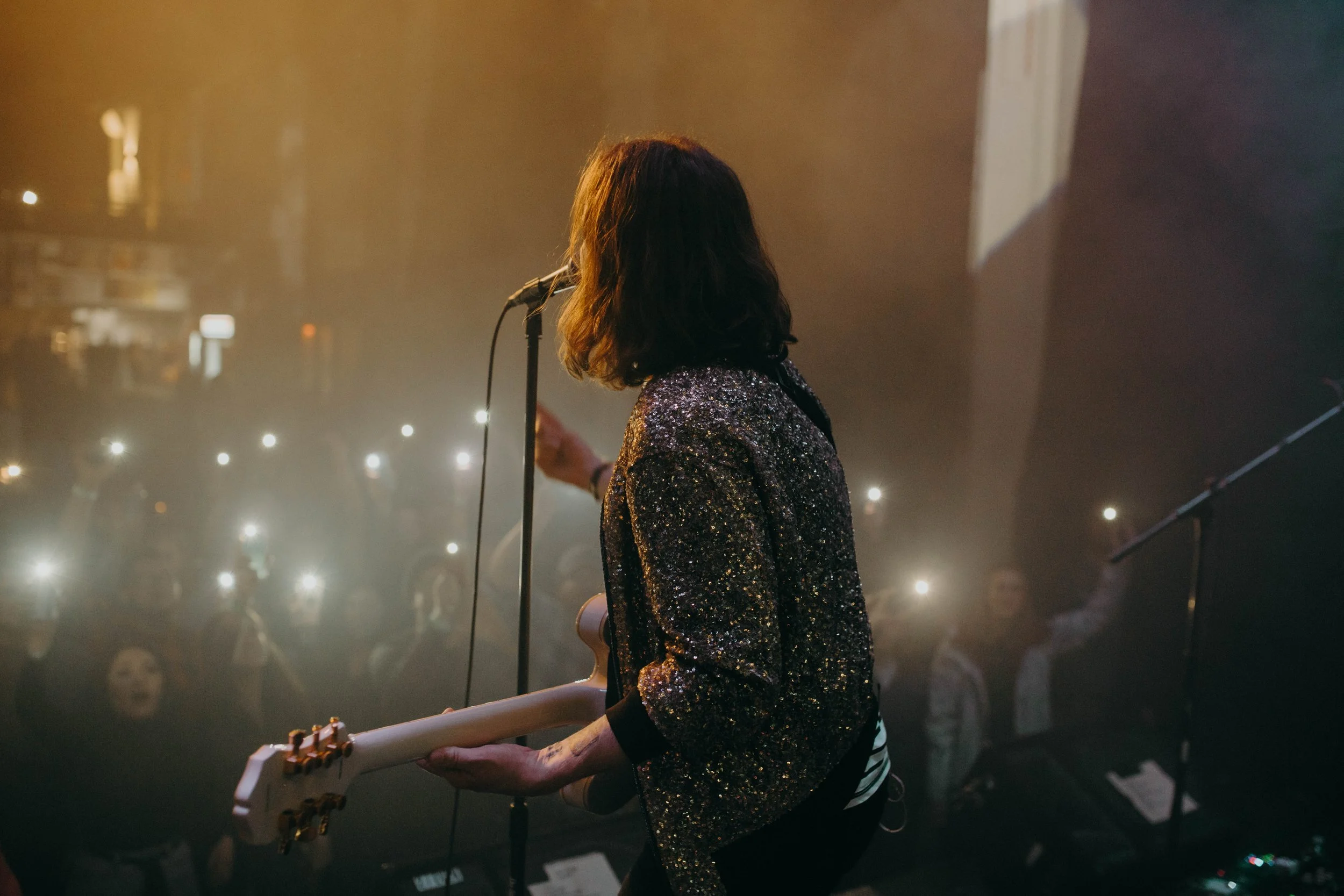 A musician, possibly a female, with shoulder-length dark hair, performing on stage with a guitar and dressed in a sparkly jacket, facing an audience with many fans holding up their phones with flashlights on.