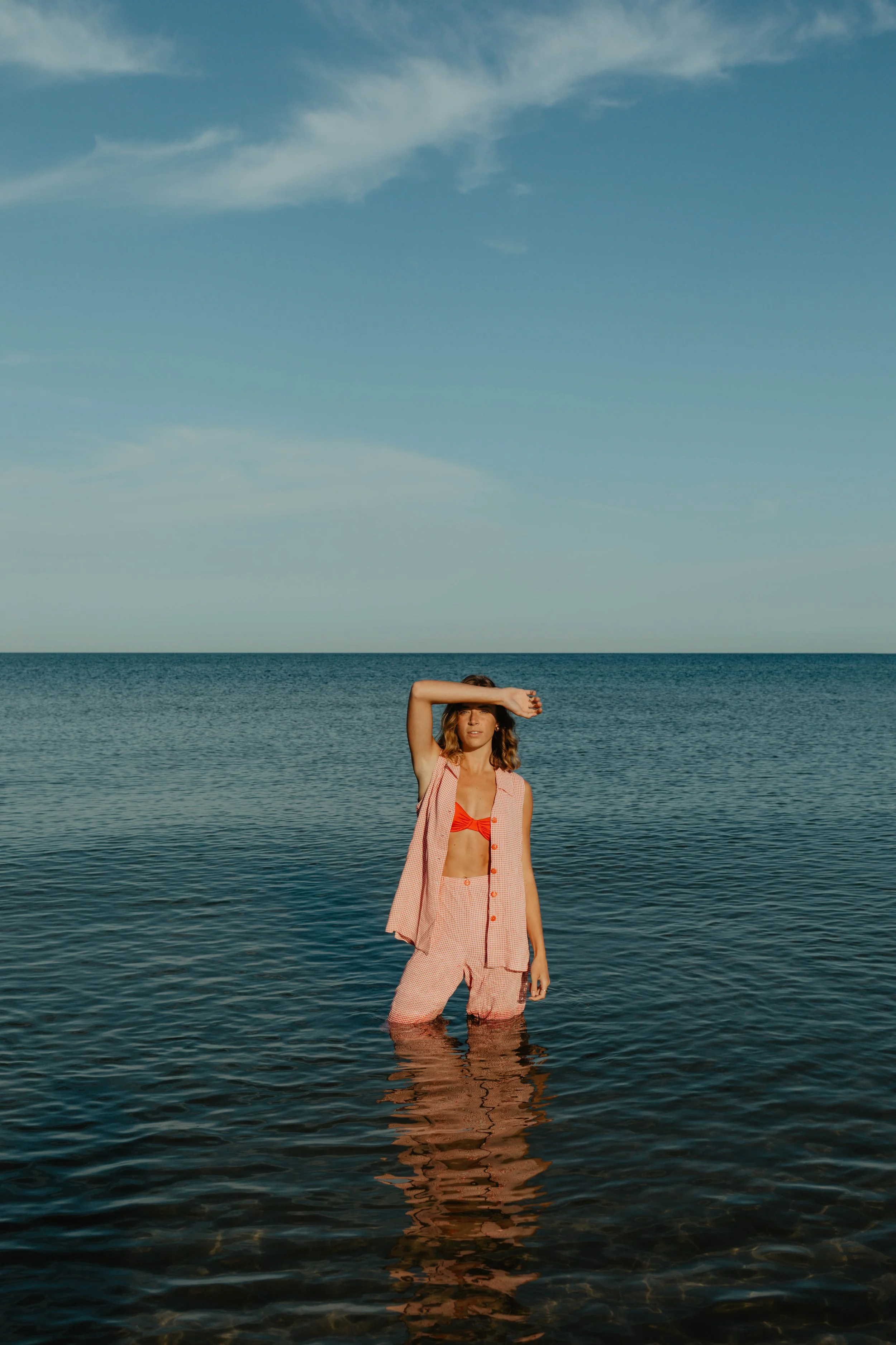 A woman standing in the ocean with her arm raised across her forehead, wearing a pink sleeveless button-up shirt and matching shorts, with a red bikini top underneath, under a blue sky.