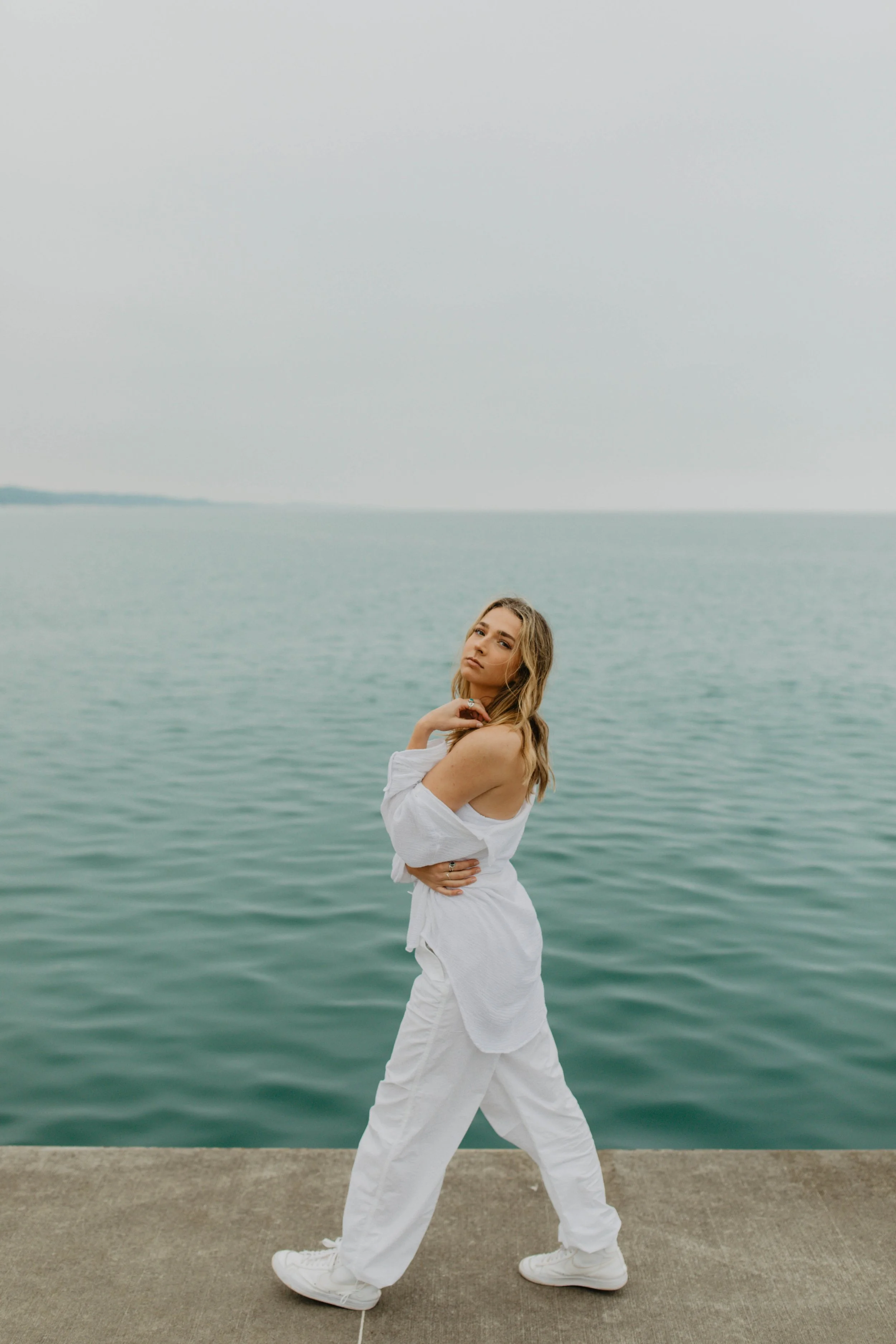 A young woman in white clothing standing on a concrete pier near a body of water, with an overcast sky in the background.