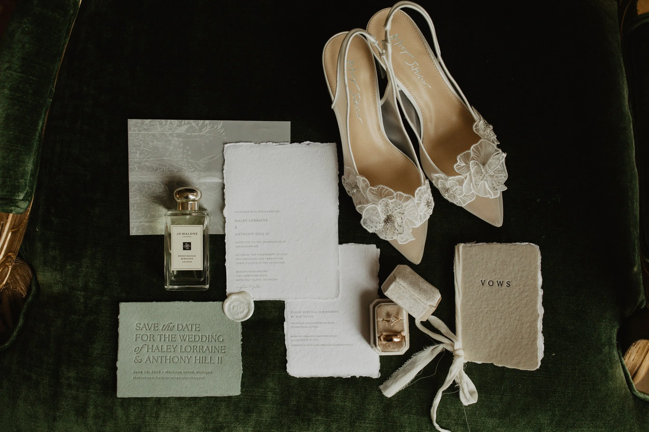 Wedding invitation, perfume bottle, pair of beige high heels with floral details, jewelry box with rings, wedding ring on a ring pillow, 'Save the Date' card, and a book titled 'Vows', arranged on a dark green surface.