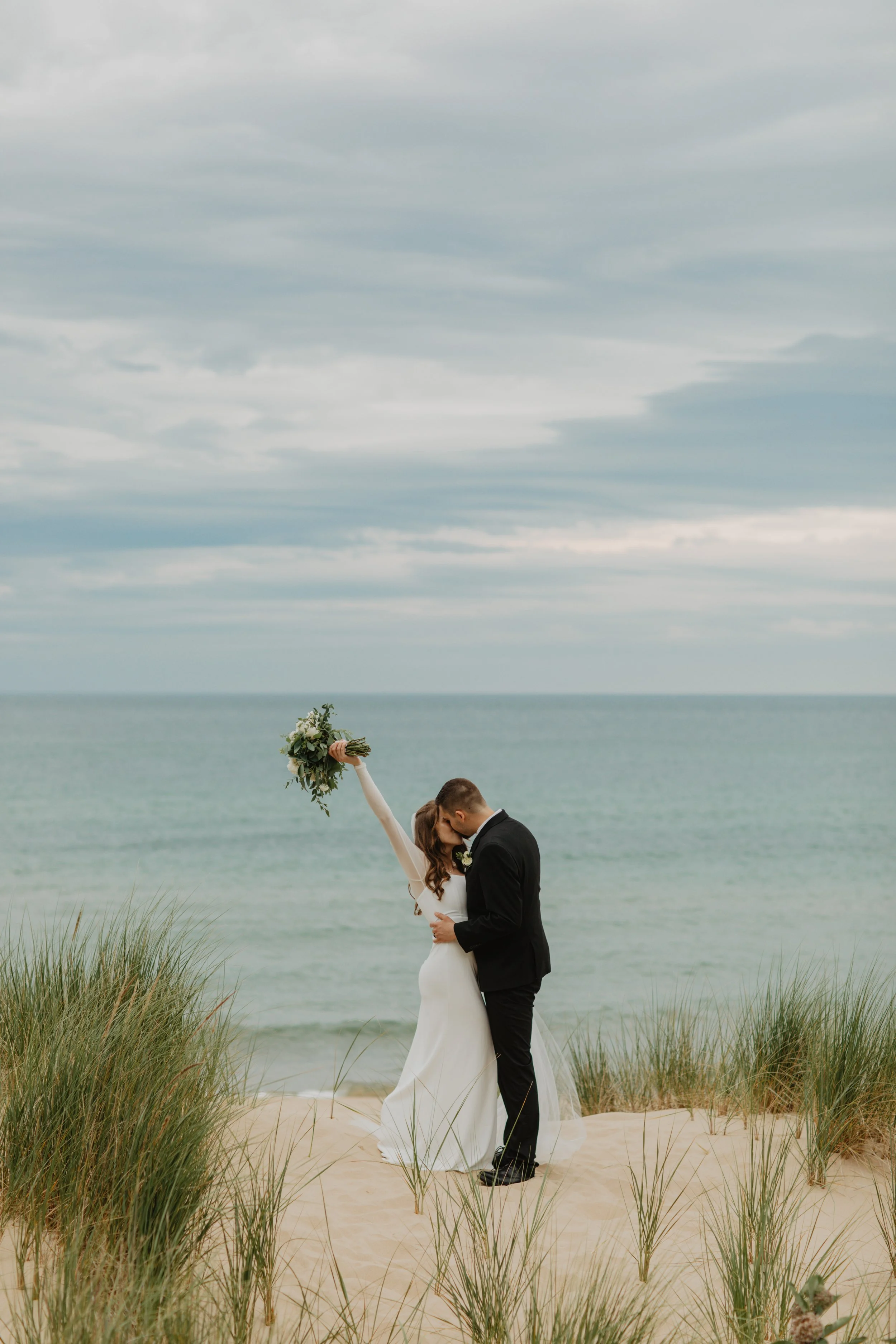 A bride and groom kissing on a sandy beach with grass, ocean, and cloudy sky in the background. The bride is wearing a white wedding dress and holding a bouquet, while the groom is dressed in a black suit.