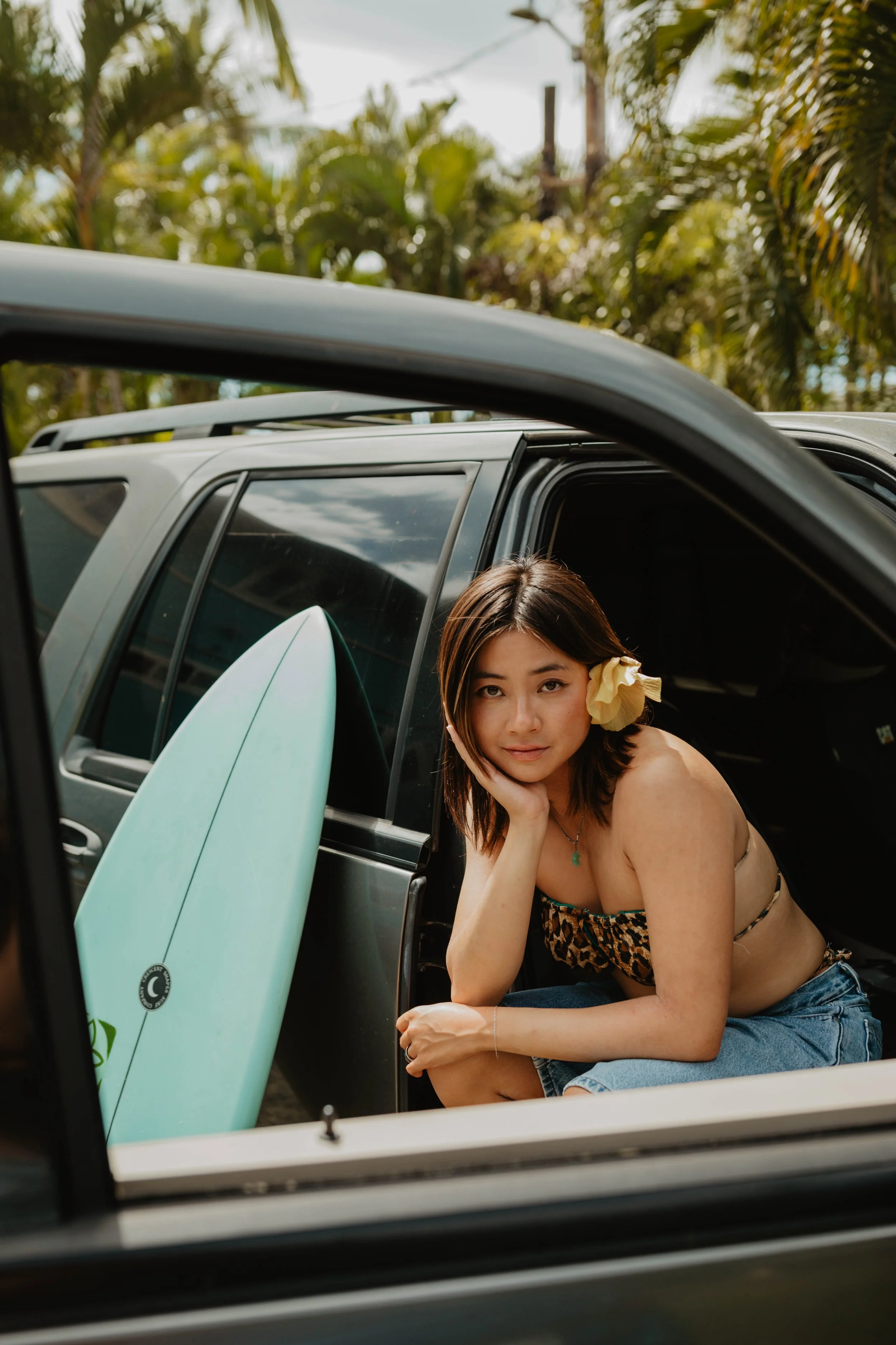 A woman with a flower in her hair sitting in the open door of a black SUV, with a surfboard leaning against the door, surrounded by tropical trees.