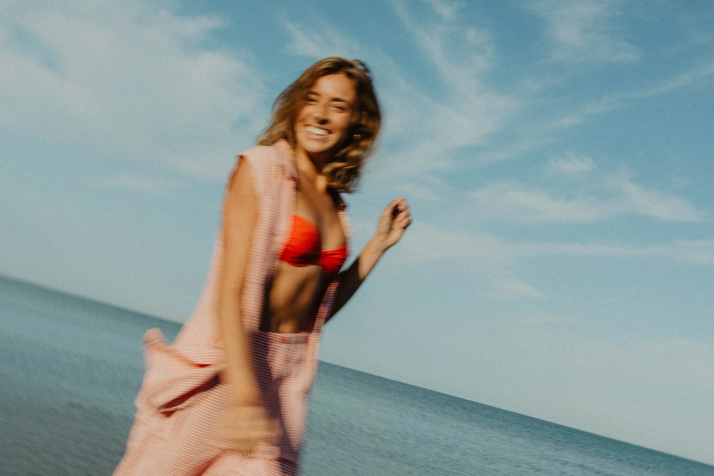 A young woman in a red swimsuit and pink cover-up smiling and running on a beach with a blue sky and ocean in the background.