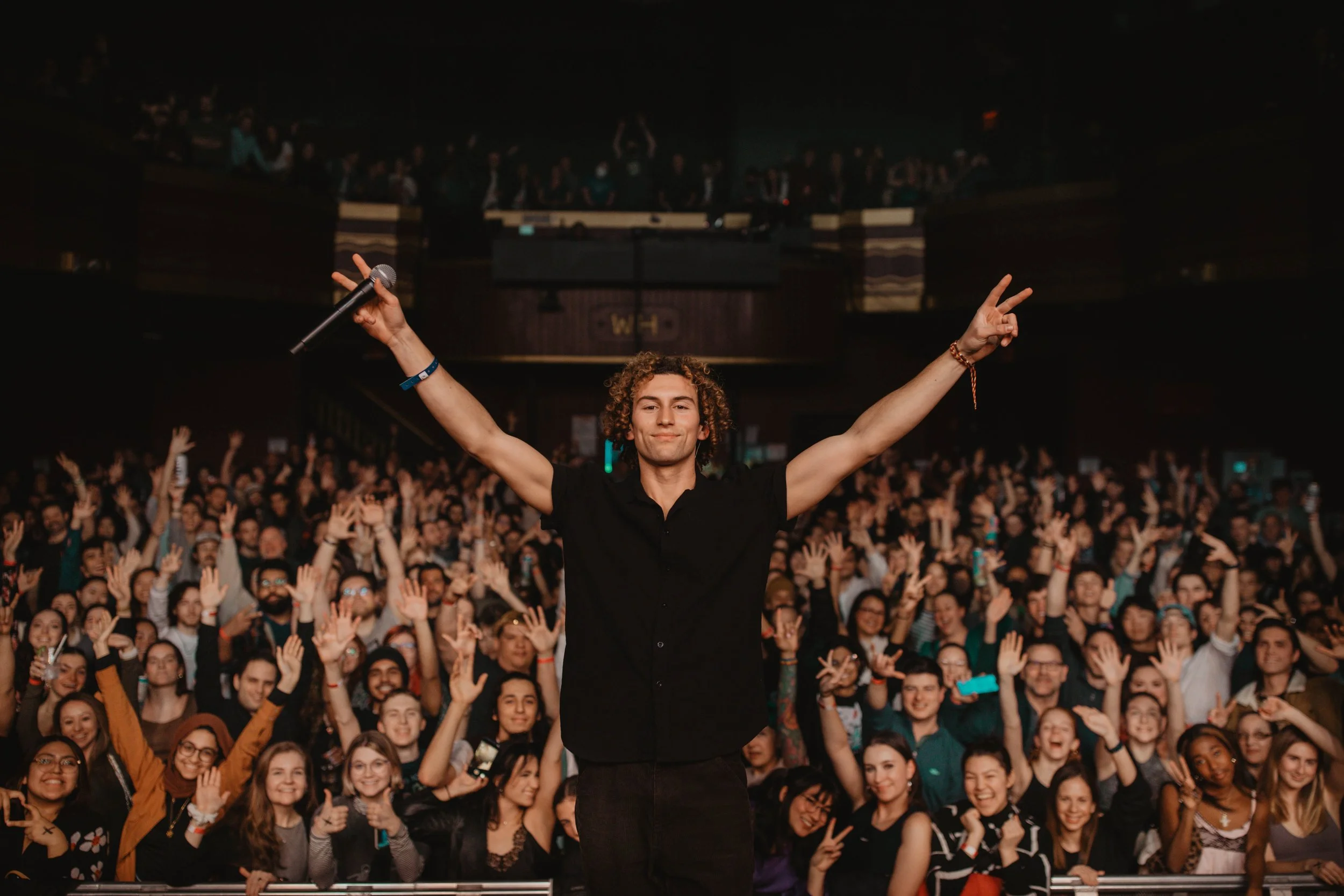 The music artist Sammy Rash in a black shirt on stage with arms raised, holding a microphone in one hand, in front of a cheering crowd at a concert.