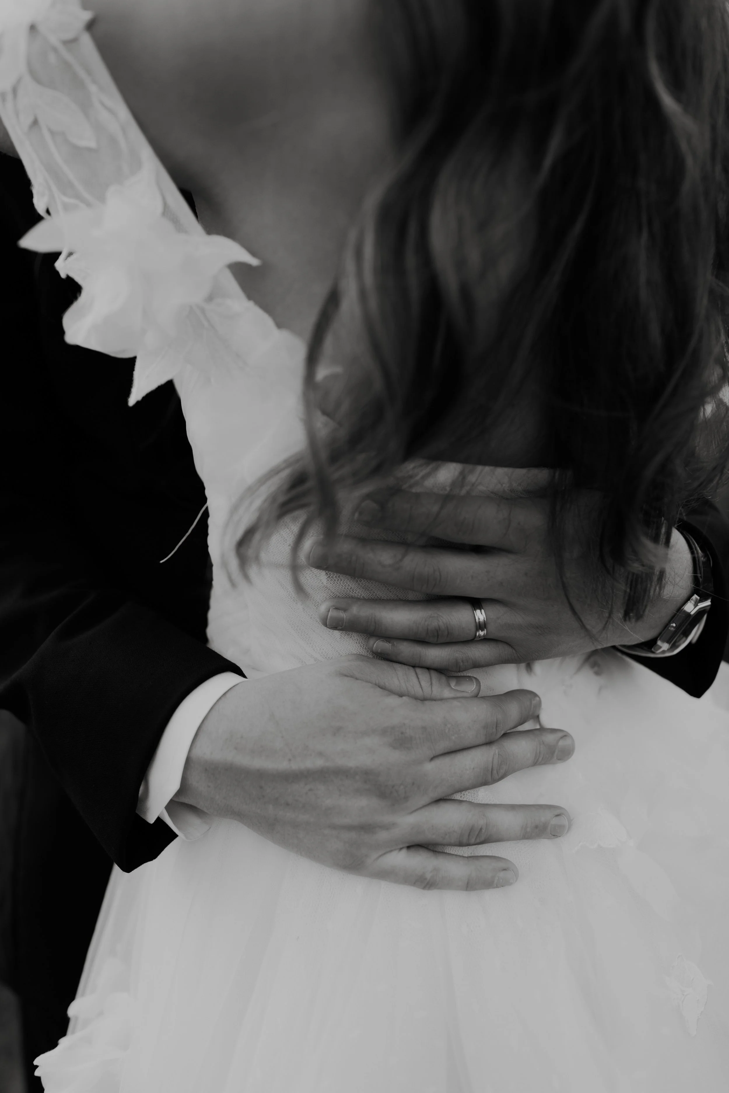 A close-up of a couple holding each other during a wedding, with the bride and groom's hands and part of the woman's dress visible.