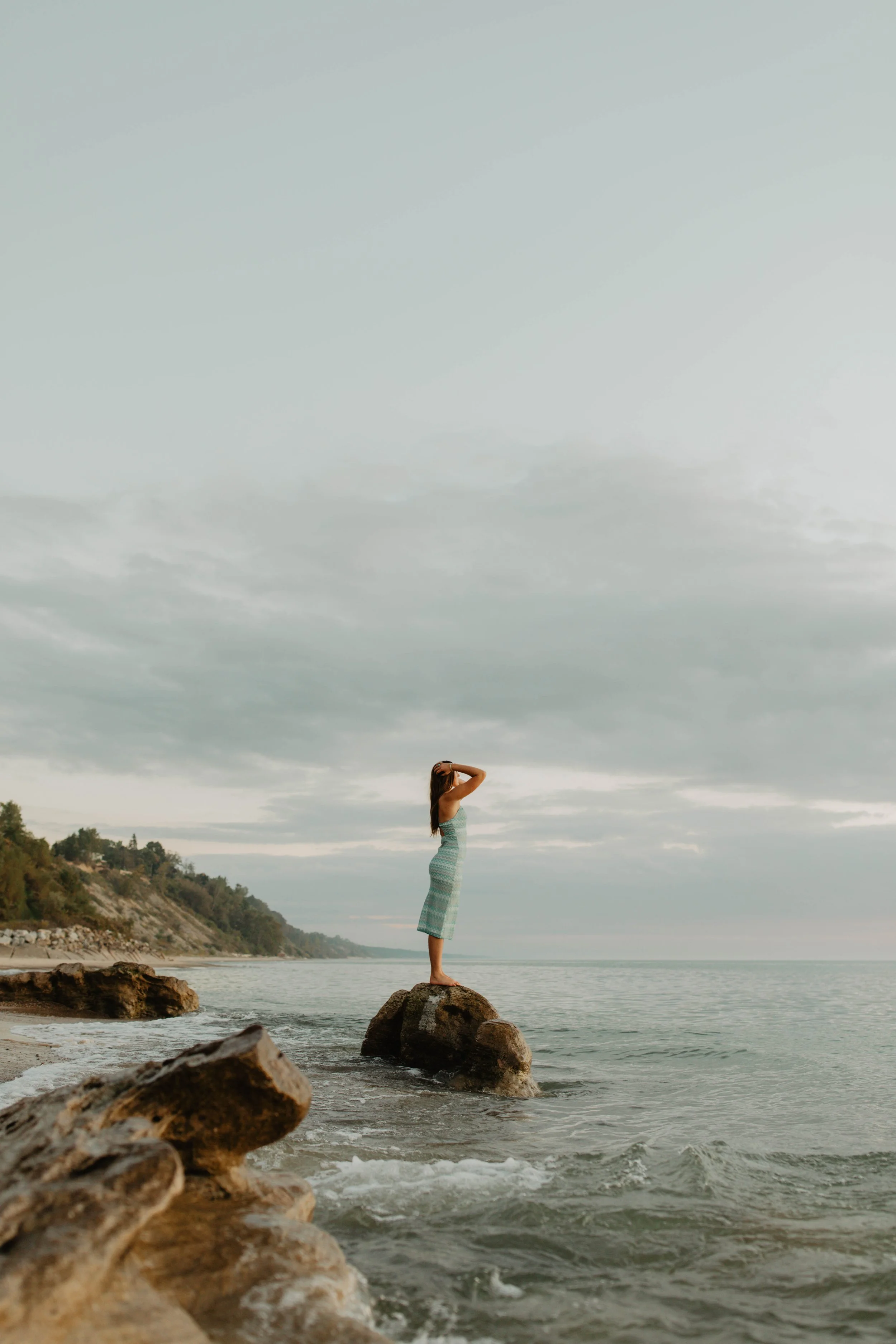 A woman stands on a rock at the shoreline, facing the ocean with her back to the camera, during cloudy weather.