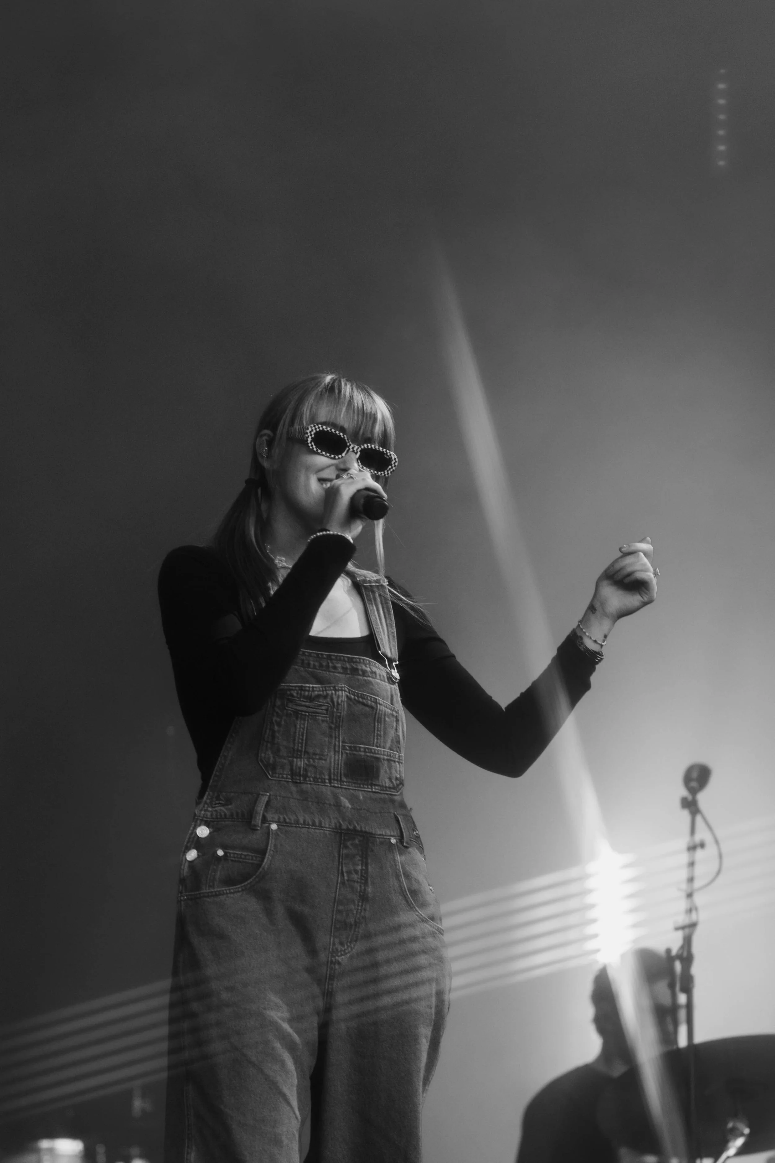 Sasha Sloan performing on stage with a microphone, wearing sunglasses and overalls, with stage lighting and a microphone stand in the background in Detroit, Michigan.