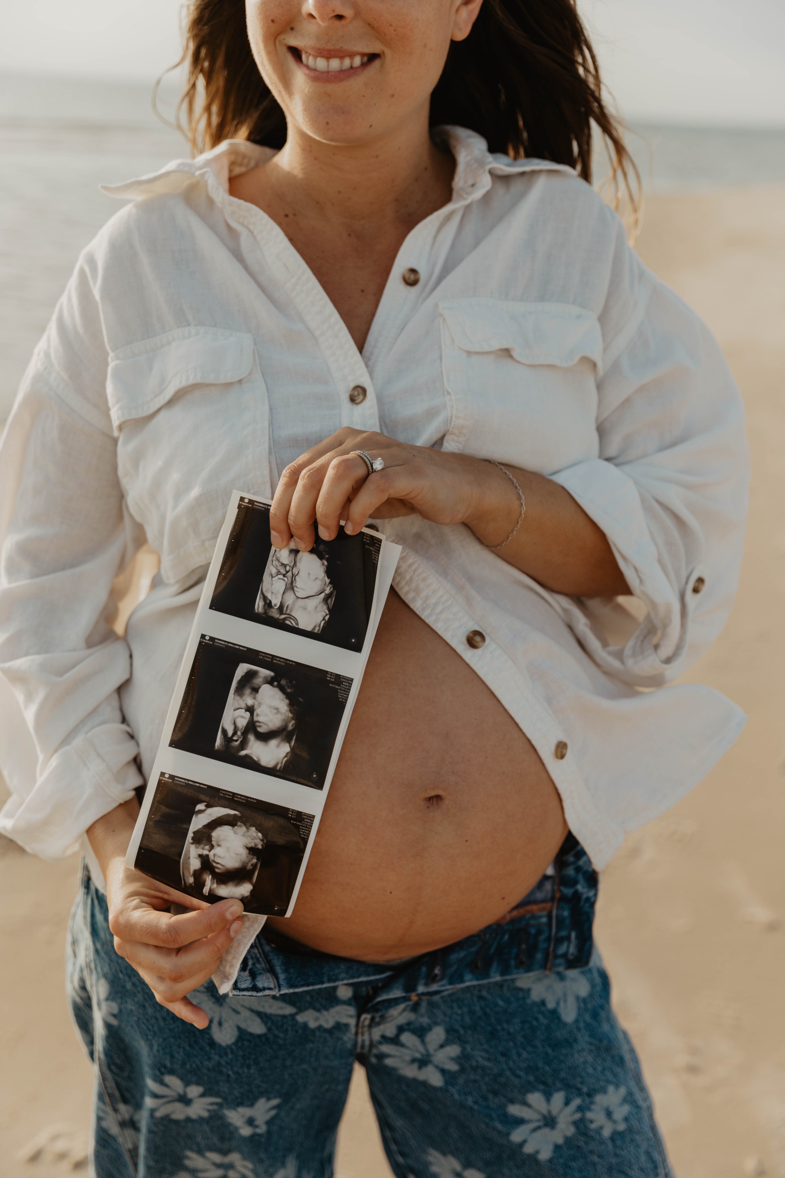 Pregnant woman at the beach holding ultrasound images near her belly and smiling.