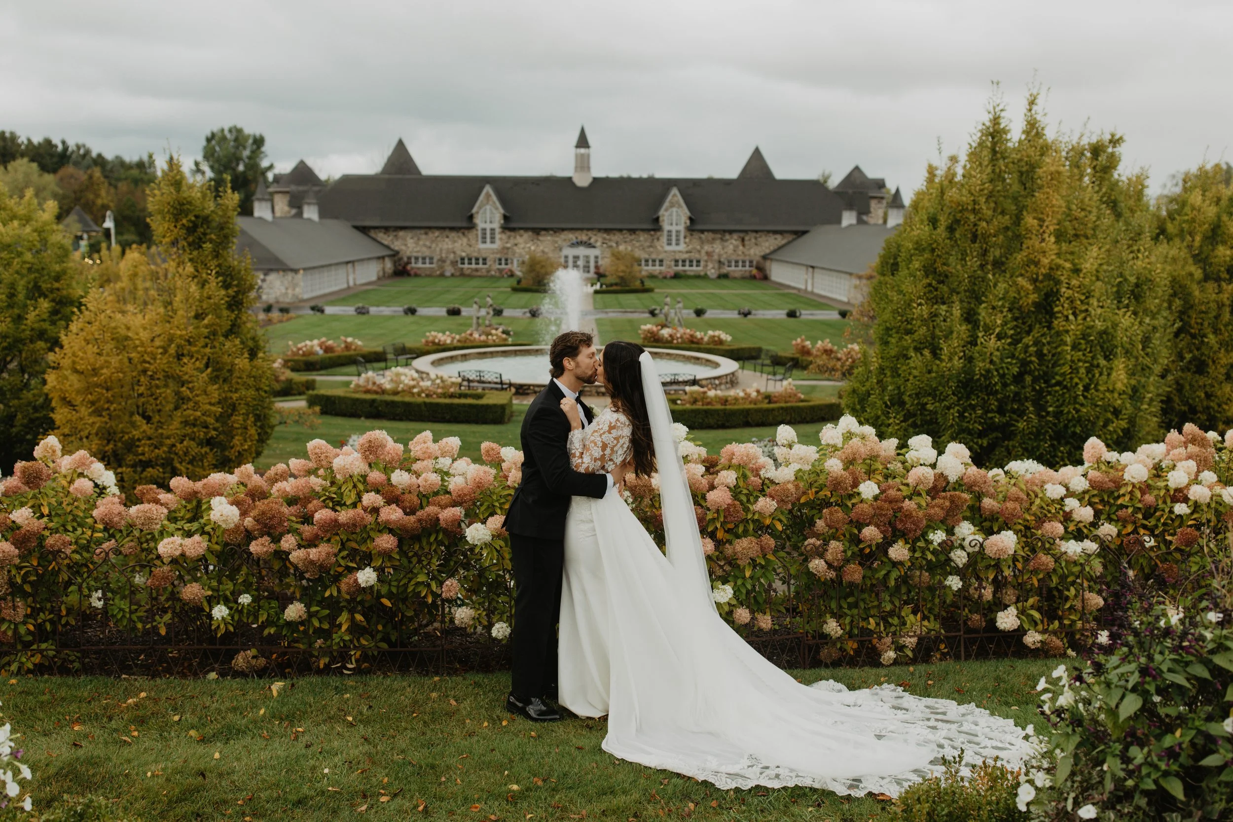 A bride and groom share a kiss in front of a garden with pink and white hydrangeas, with a large, stone mansion and fountain in the background.