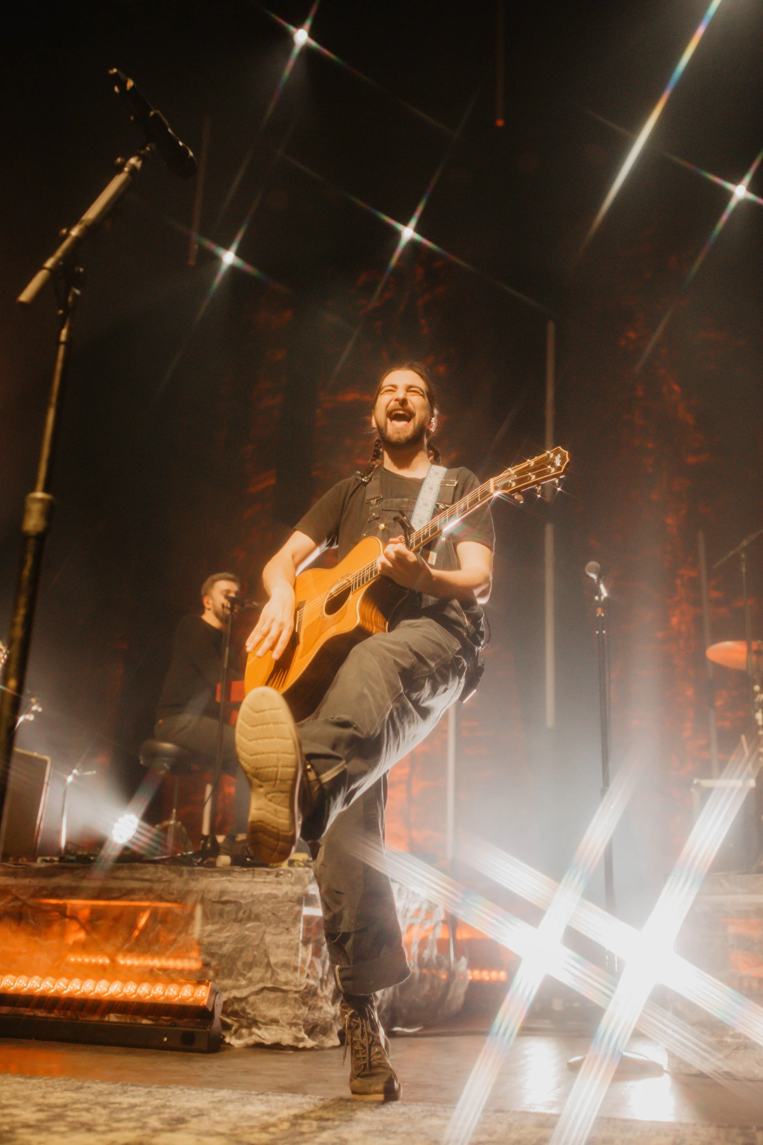 Noah Kahan playing acoustic guitar on stage, smiling with his foot raised, with bright stage lights in Detroit Michigan.