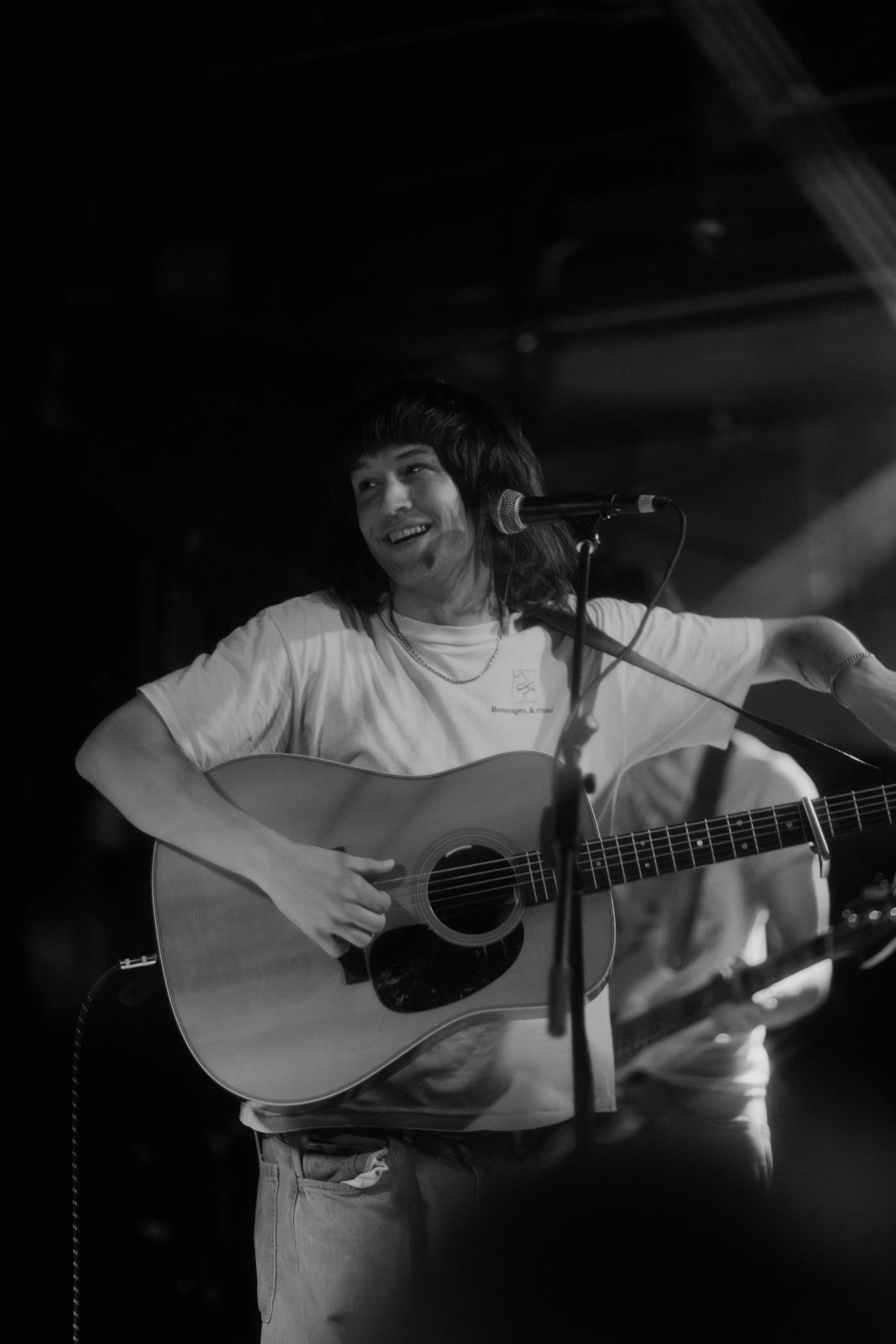 Black and white photo of the musician Dipsea Flower smiling while playing an acoustic guitar on stage, with a microphone in front of them.