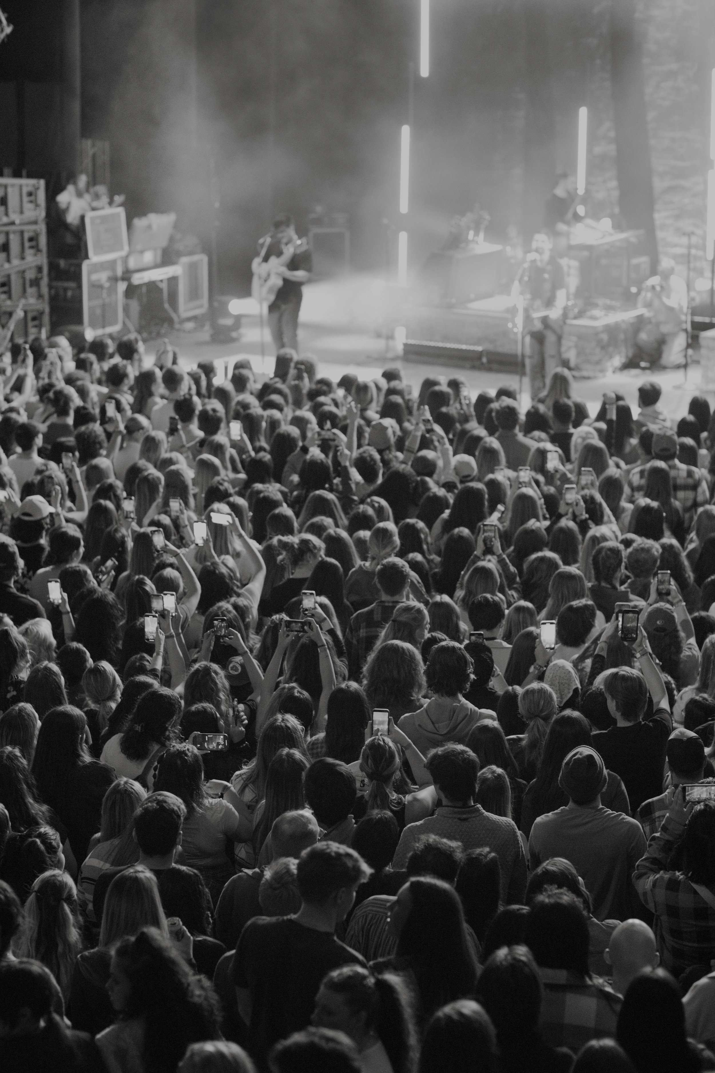 Black and white photo of a large crowd at a Noah Kahan concert, with many people holding up smartphones to record the performance on stage, where a band is playing in Detroit, Michigan.
