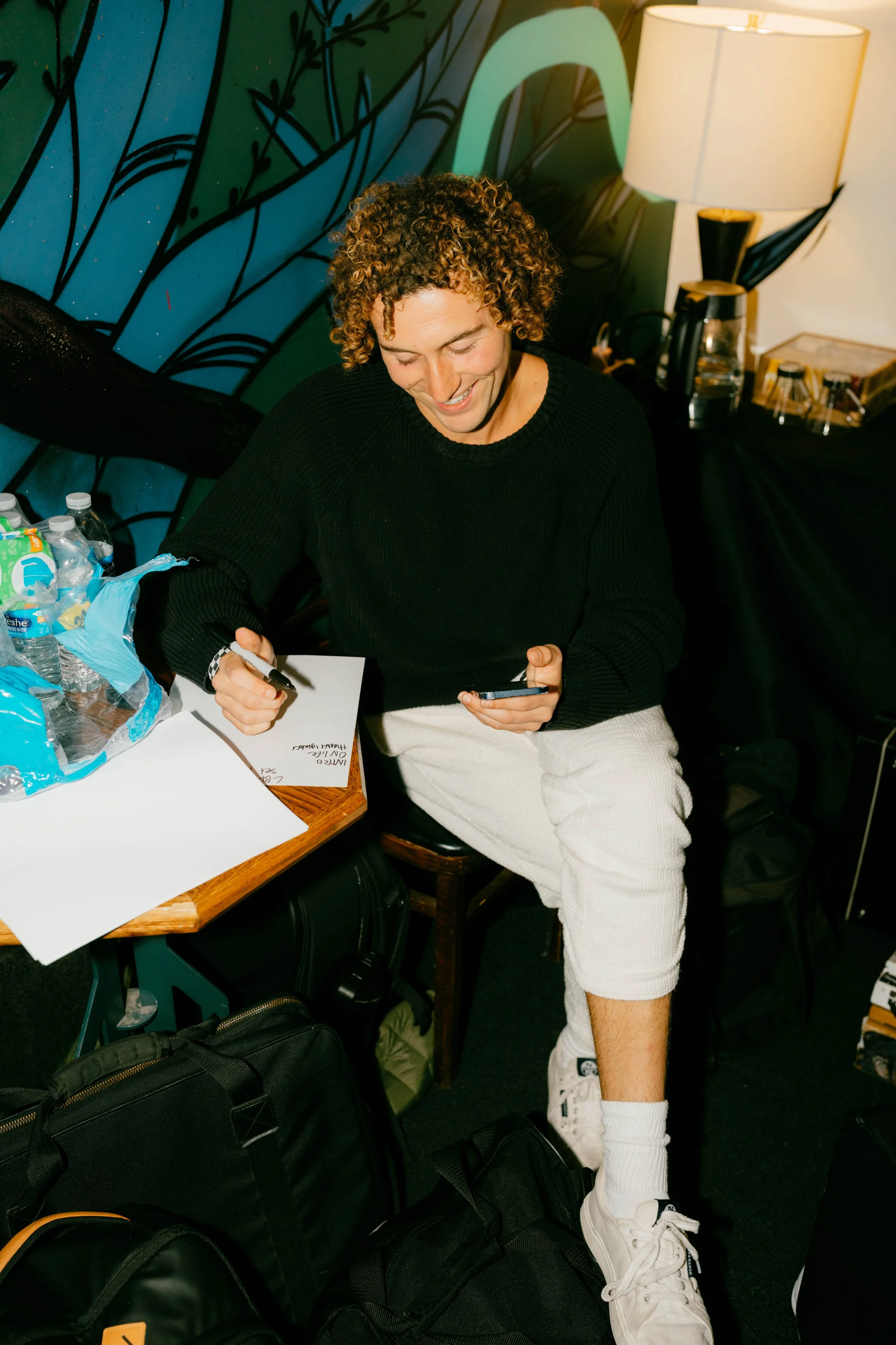 The music artist, Sammy Rash, is sitting at a table, smiling, looking at their phone, with water bottles and papers on the table, and luggage at their feet.