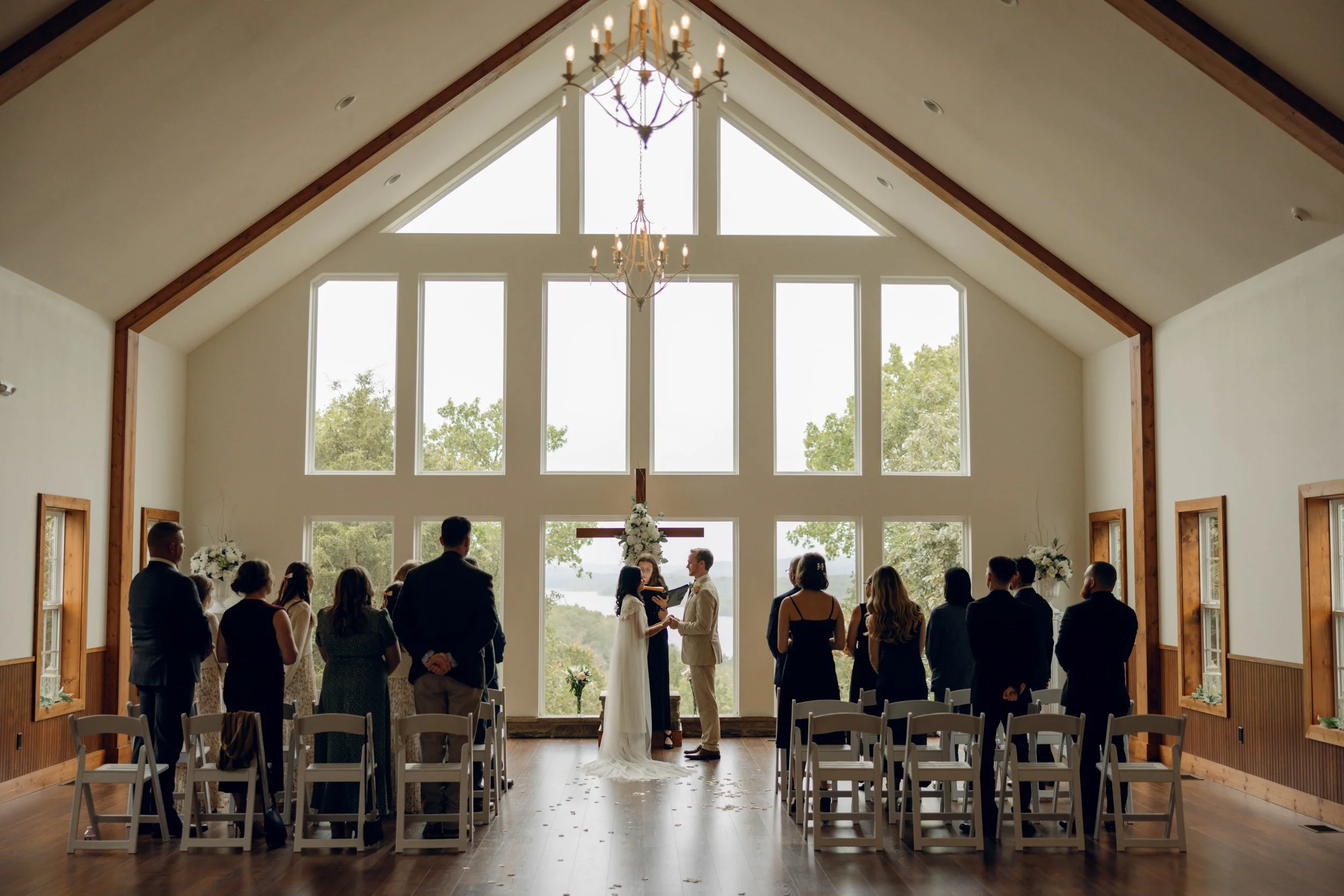 Indoor wedding ceremony with a couple exchanging vows in front of an altar with cross and flowers, large windows showing trees outside, and guests standing and seated on chairs.
