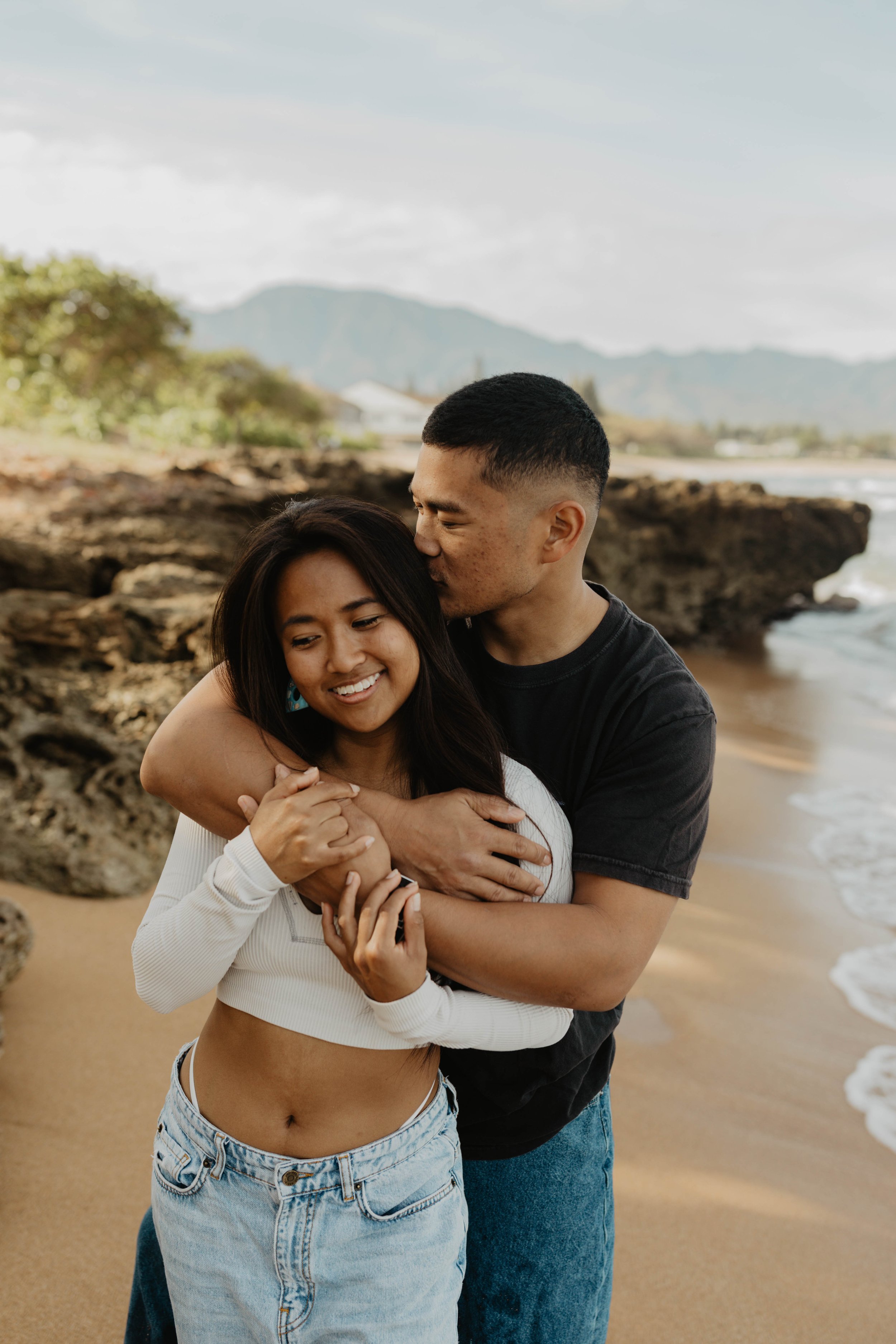 A couple embraces on a beach with rocky formations, ocean waves, and mountains in the background.