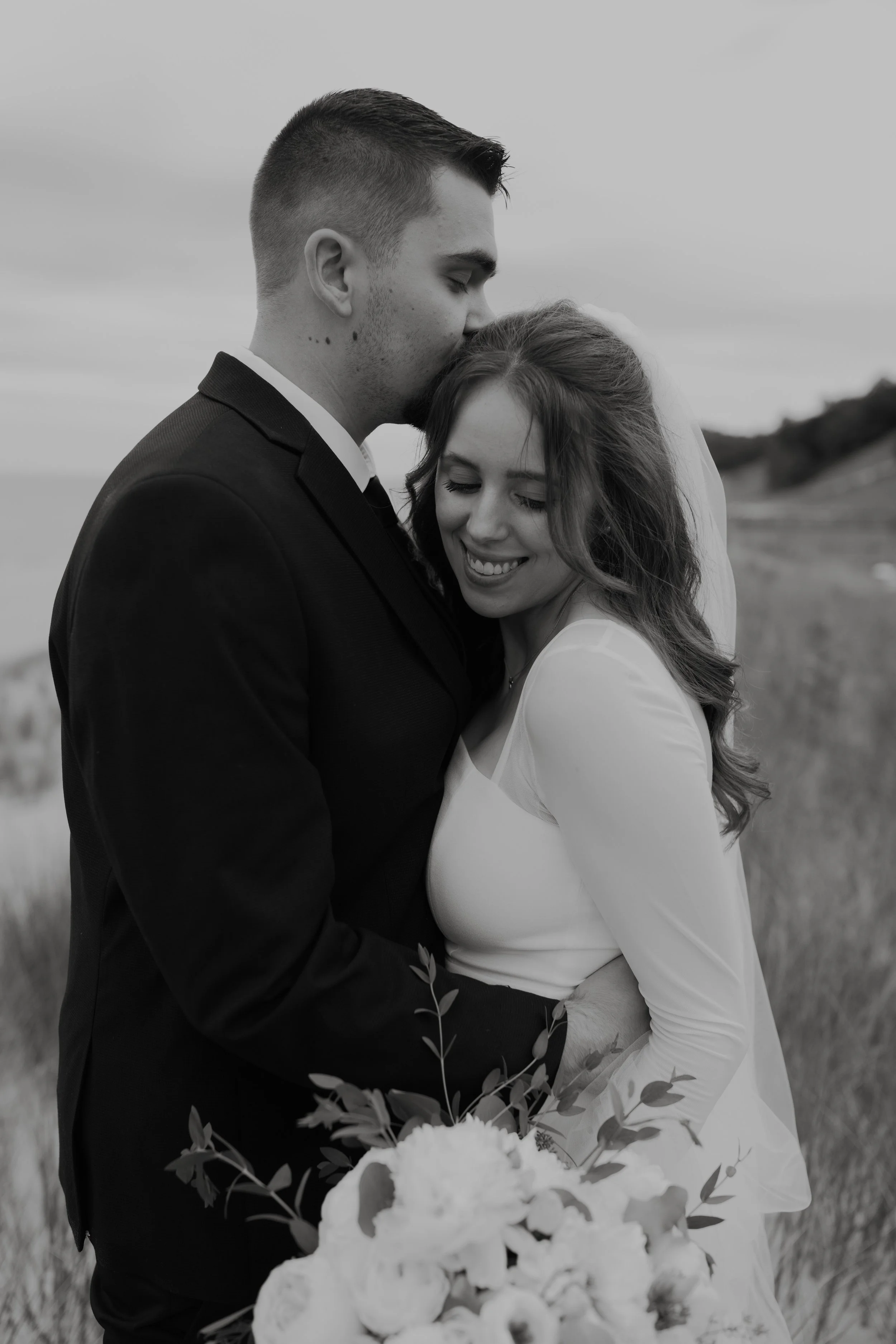 A black and white photo of a couple in wedding attire embracing outdoors, with the man kissing the woman's forehead and the woman smiling with her eyes closed, holding a bouquet of flowers.