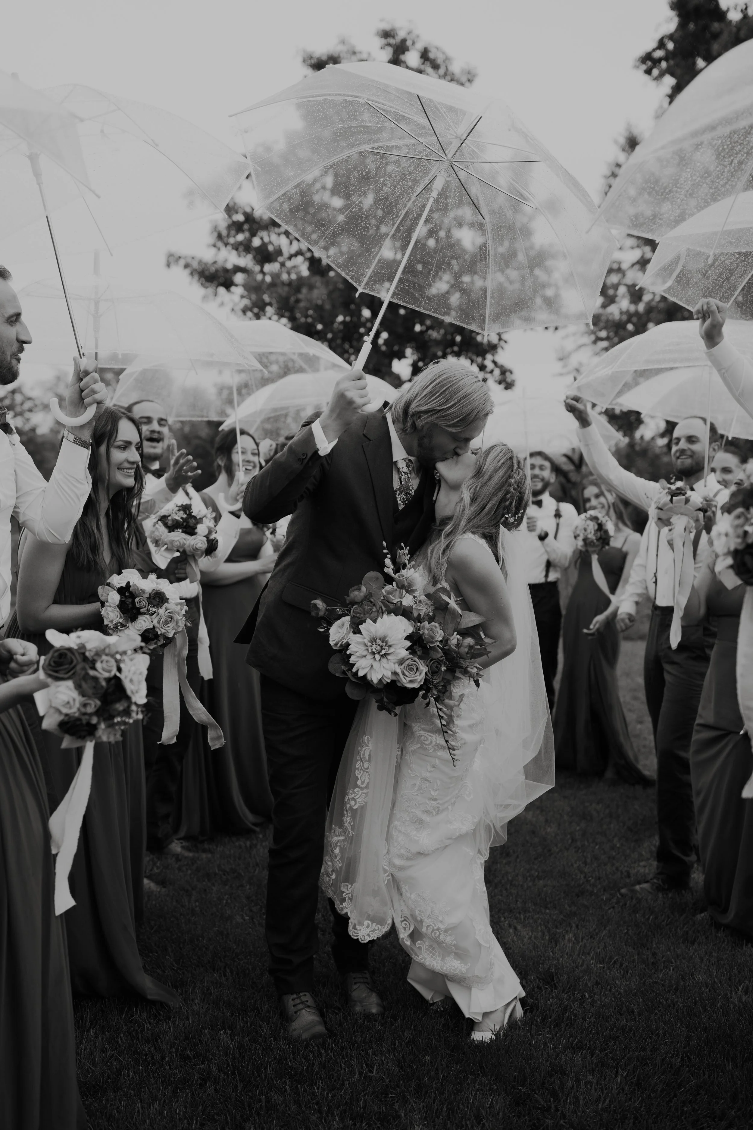 A wedding couple sharing a kiss under umbrellas with wedding guests surrounding them in a garden.