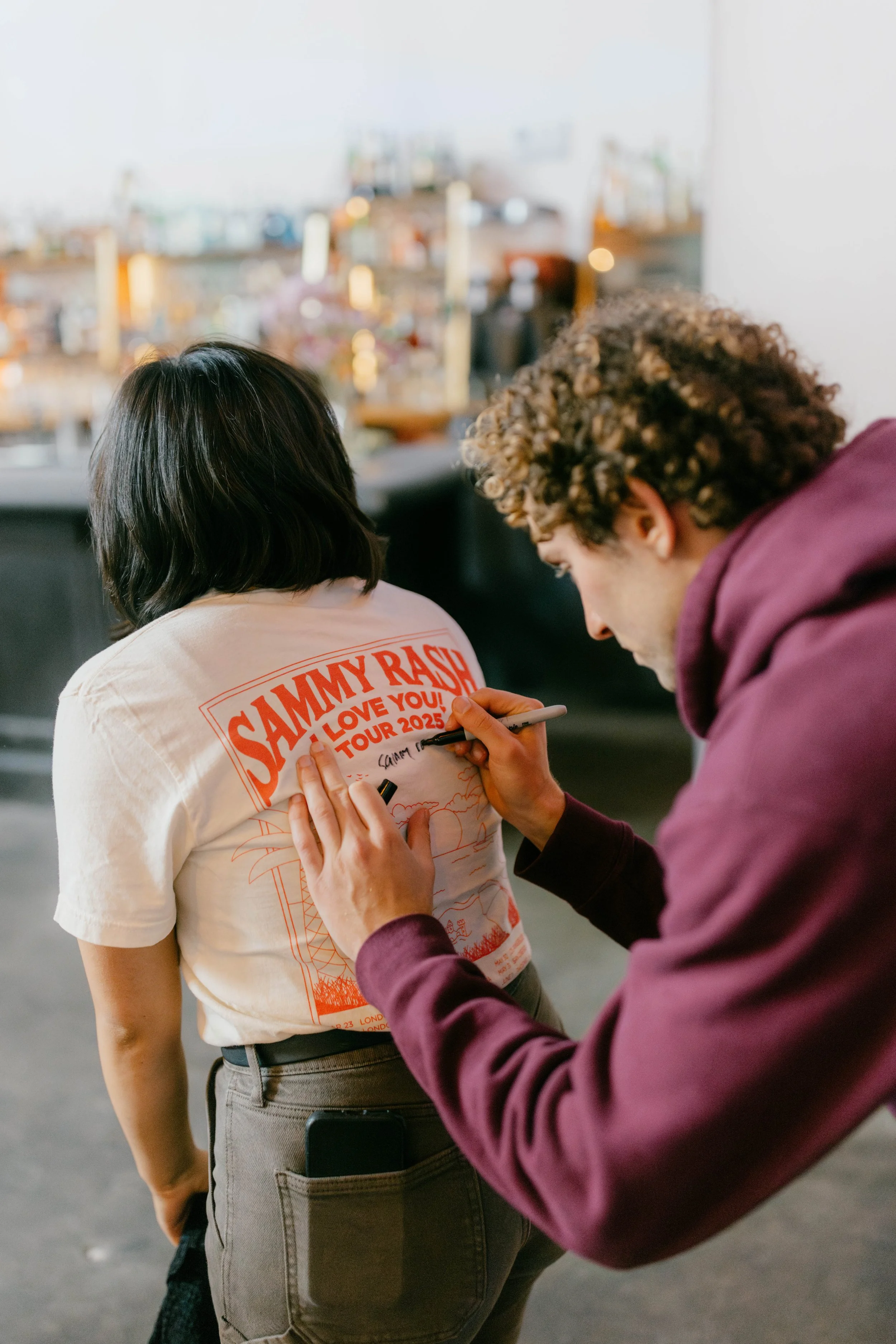 The music artist Sammy Rash  is signing the back of a woman's t-shirt that says 'SAMMY RASH TOUR 2025' in orange letters, in a casual indoor setting.
