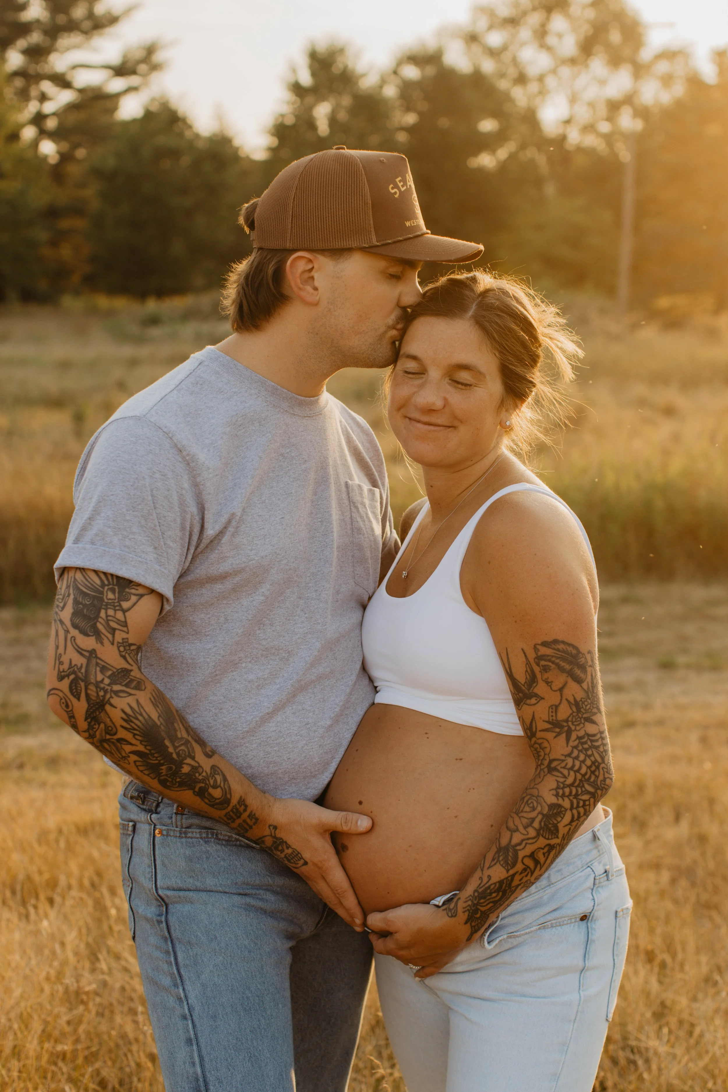 A pregnant woman with tattoos on her arms, standing outdoors in a field at sunset, smiling with her eyes closed. A man, also tattooed, is kissing her forehead while holding her baby bump, in a tender moment of affection.