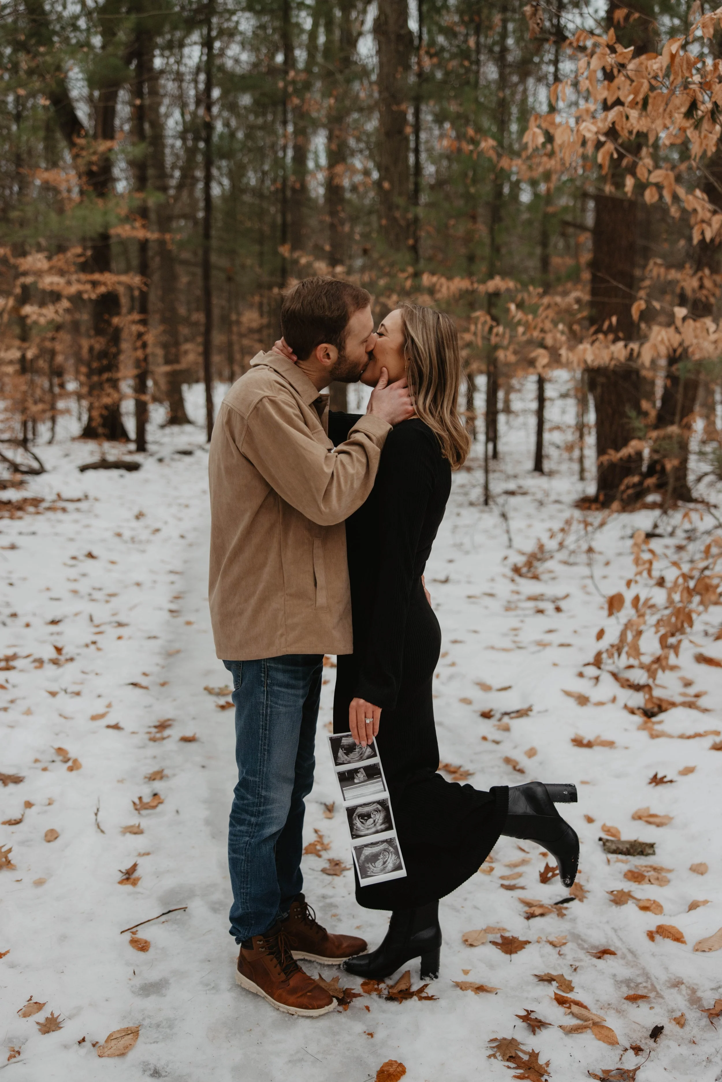 A couple kissing in a snowy forest, with the woman holding an ultrasound photo.