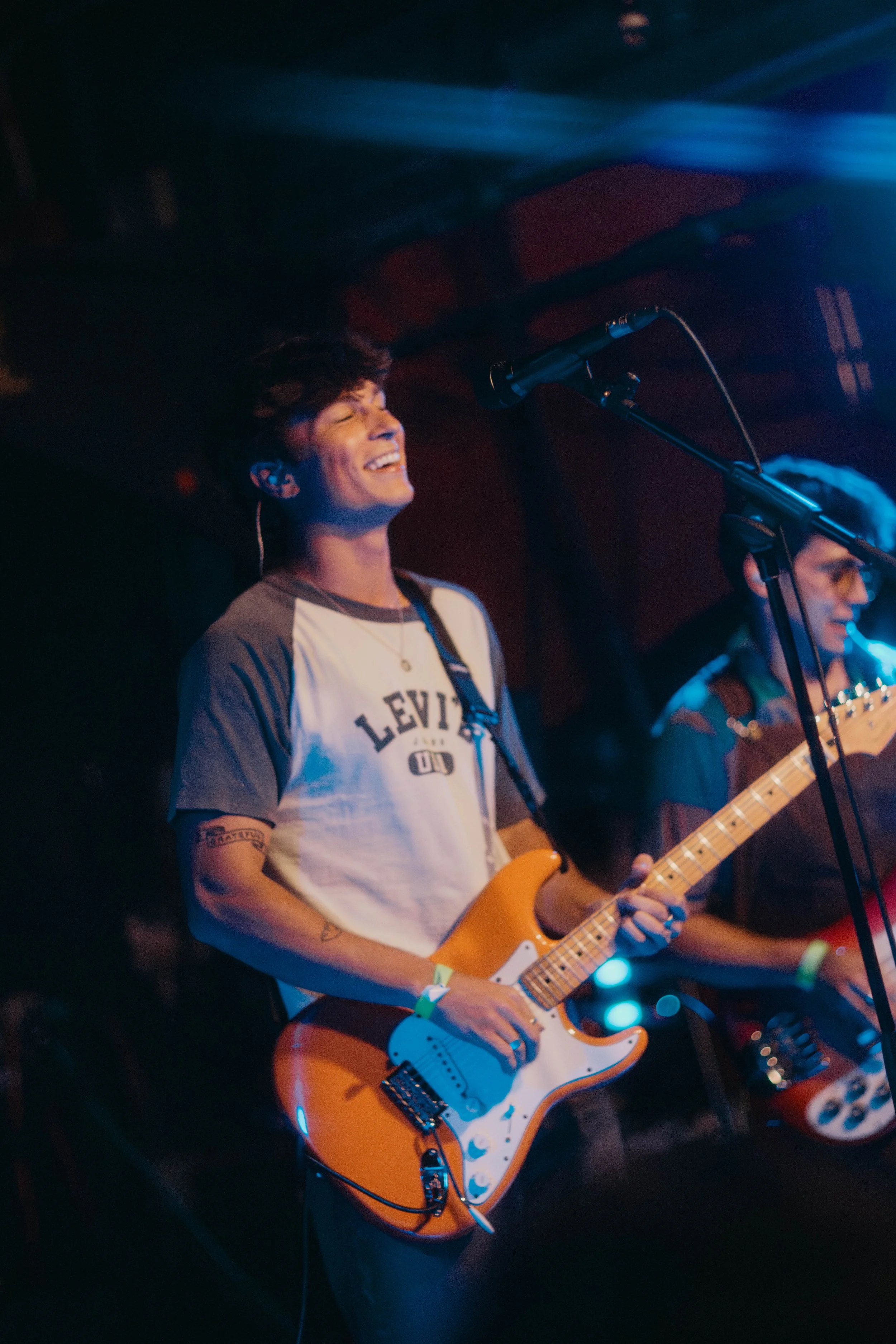 Kevin Kramer, a musician playing an electric guitar on stage, smiling with eyes closed, wearing a gray and white Levi's t-shirt, and standing under stage lights in Boston.
