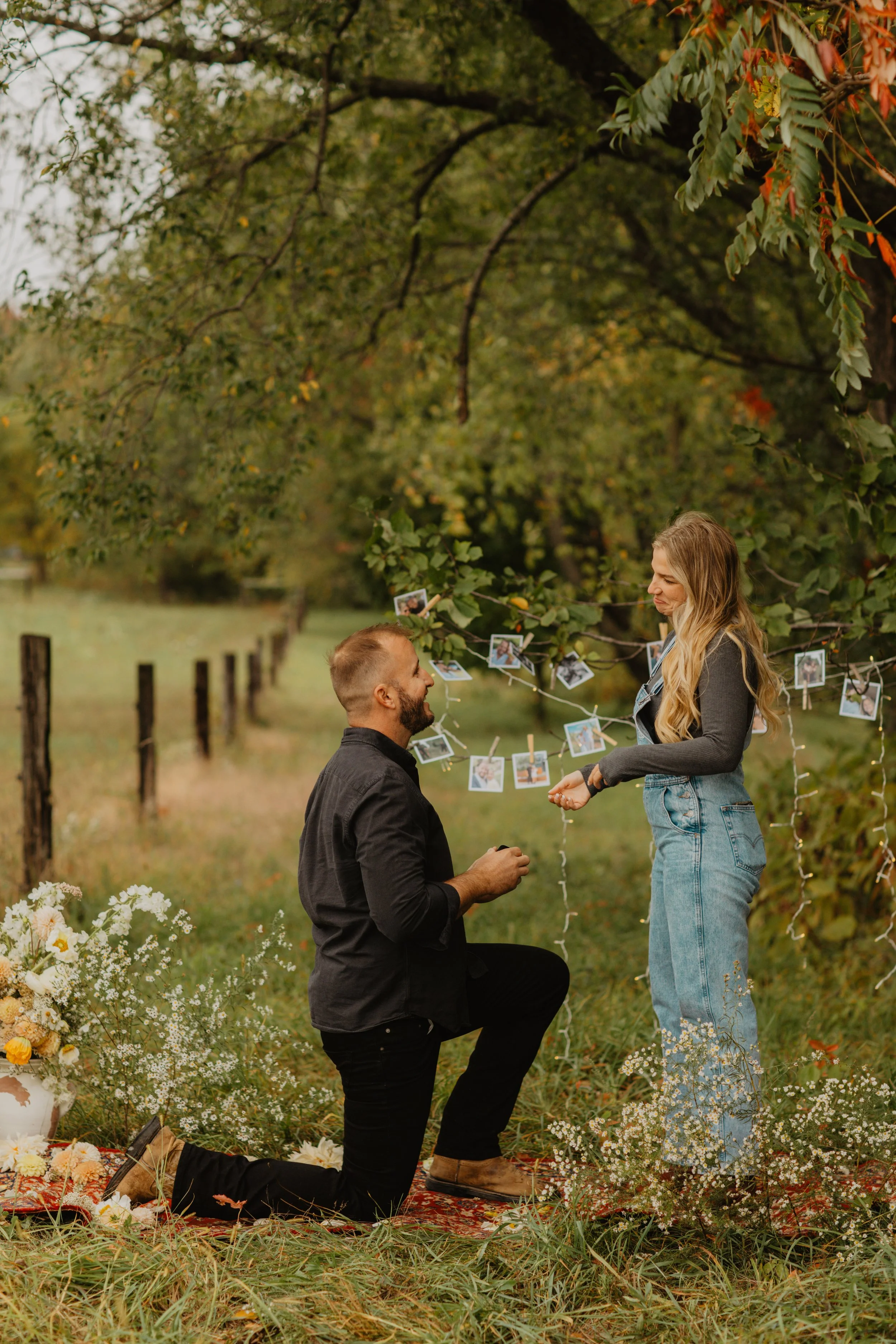 A man is kneeling on one knee proposing to a woman in a rustic outdoor setting. The woman is standing, smiling, and holding the man's hand. There are photographs hanging on a string behind them, and flowers on the ground and in a vase nearby. The sce