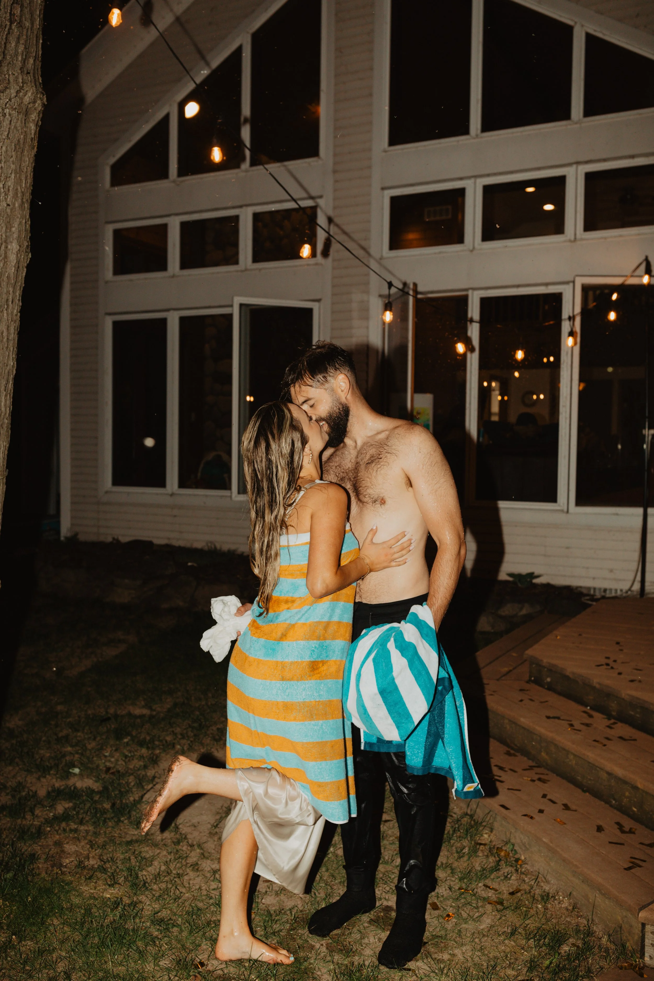 A couple is standing close together outdoors at night, about to kiss. The woman is wearing a colorful striped dress and has one leg bent backward, while the man is shirtless and holding a towel. There are string lights hanging above them and a large 