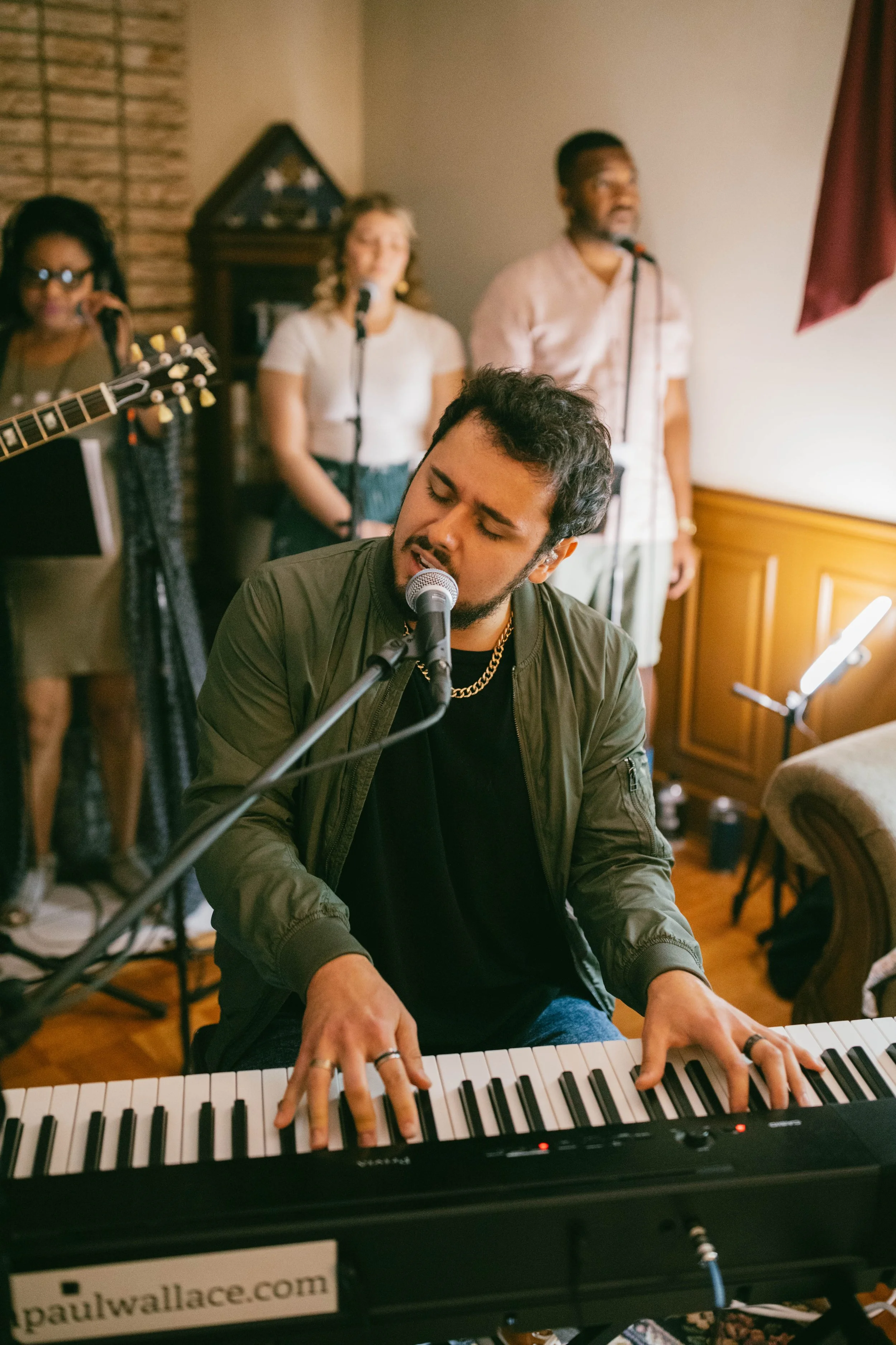 A man playing a keyboard and singing with a microphone in front of him, as a group of people sing and play guitars in the background during a live music performance.