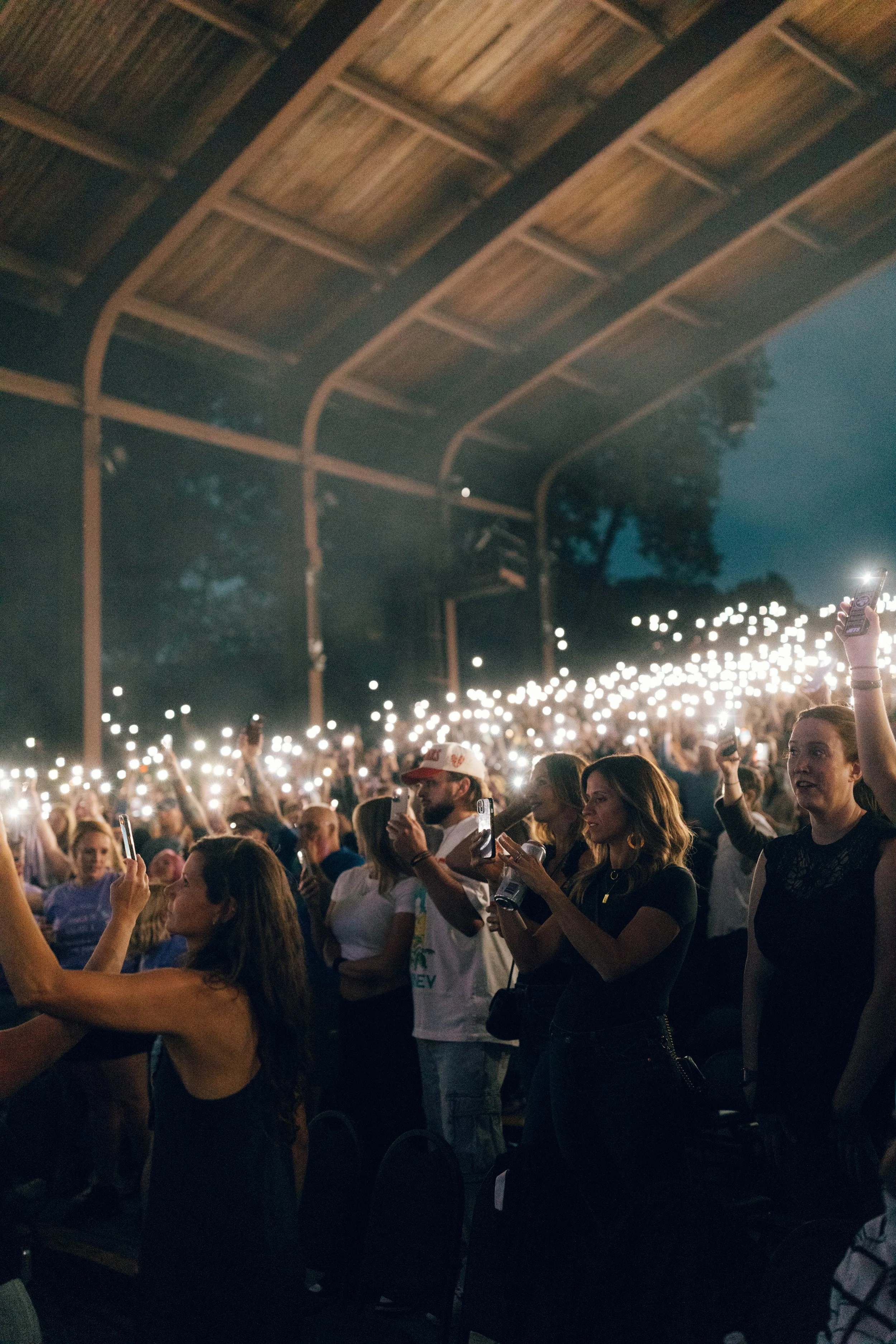 A large crowd at an outdoor concert or event at night, holding up their phones with flashlights on, under a wooden roof structure.