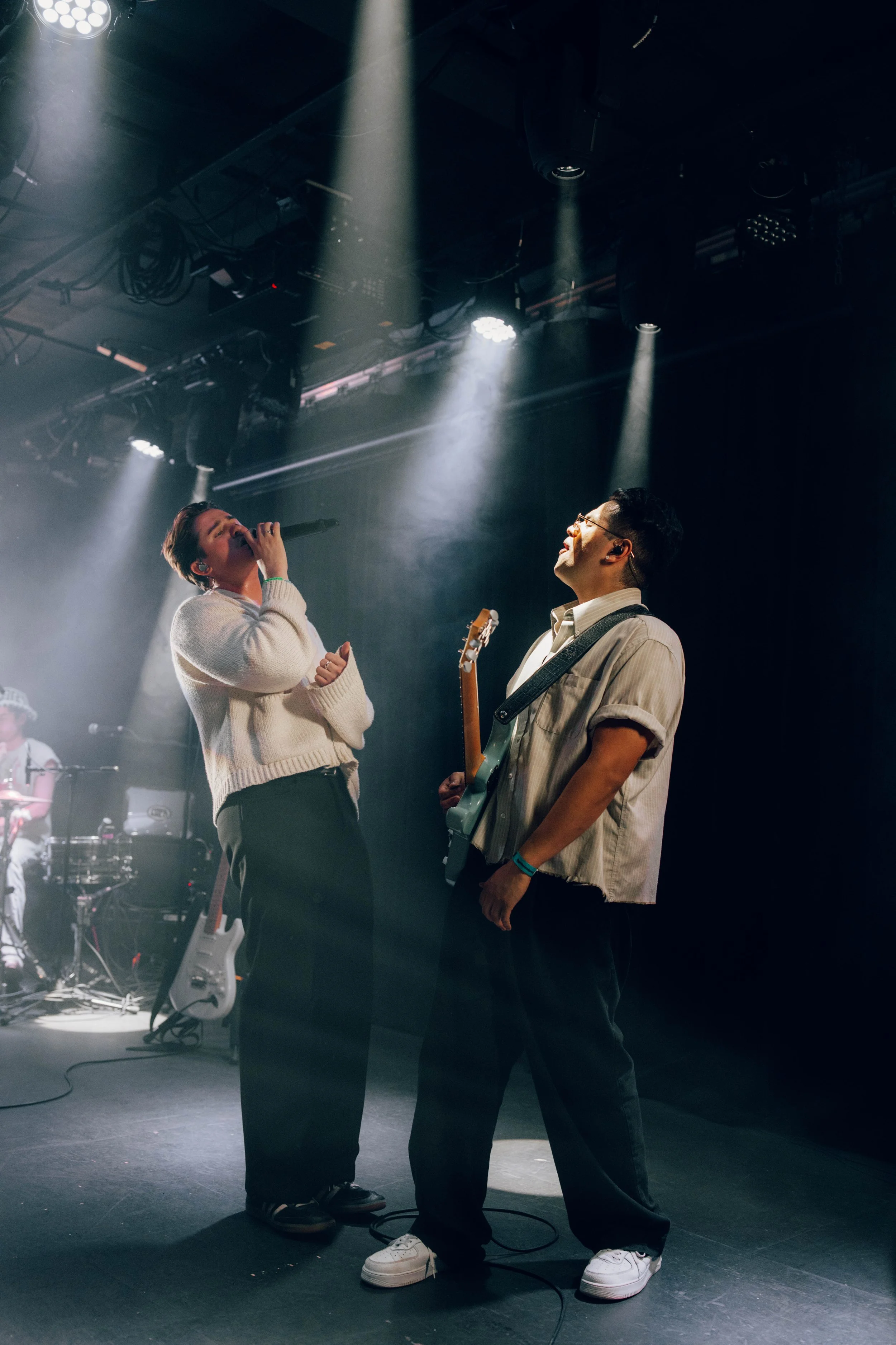 Two people performing on stage with microphones and a guitarist, under stage lights, in a concert setting.