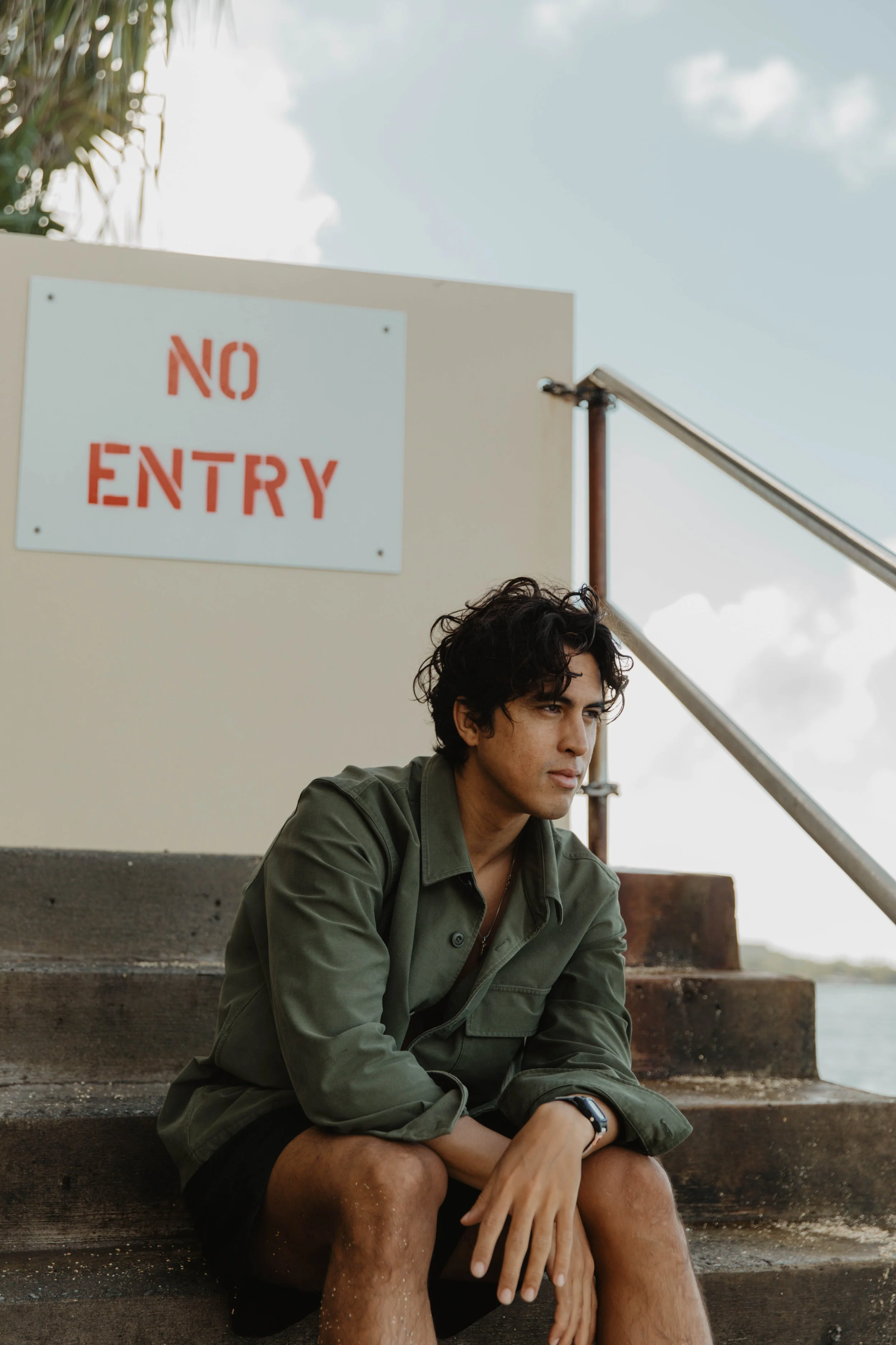 A young man with curly dark hair sitting on outdoor steps near water, wearing a green jacket and black shorts, with a serious expression, and a 'No Entry' sign visible behind him.