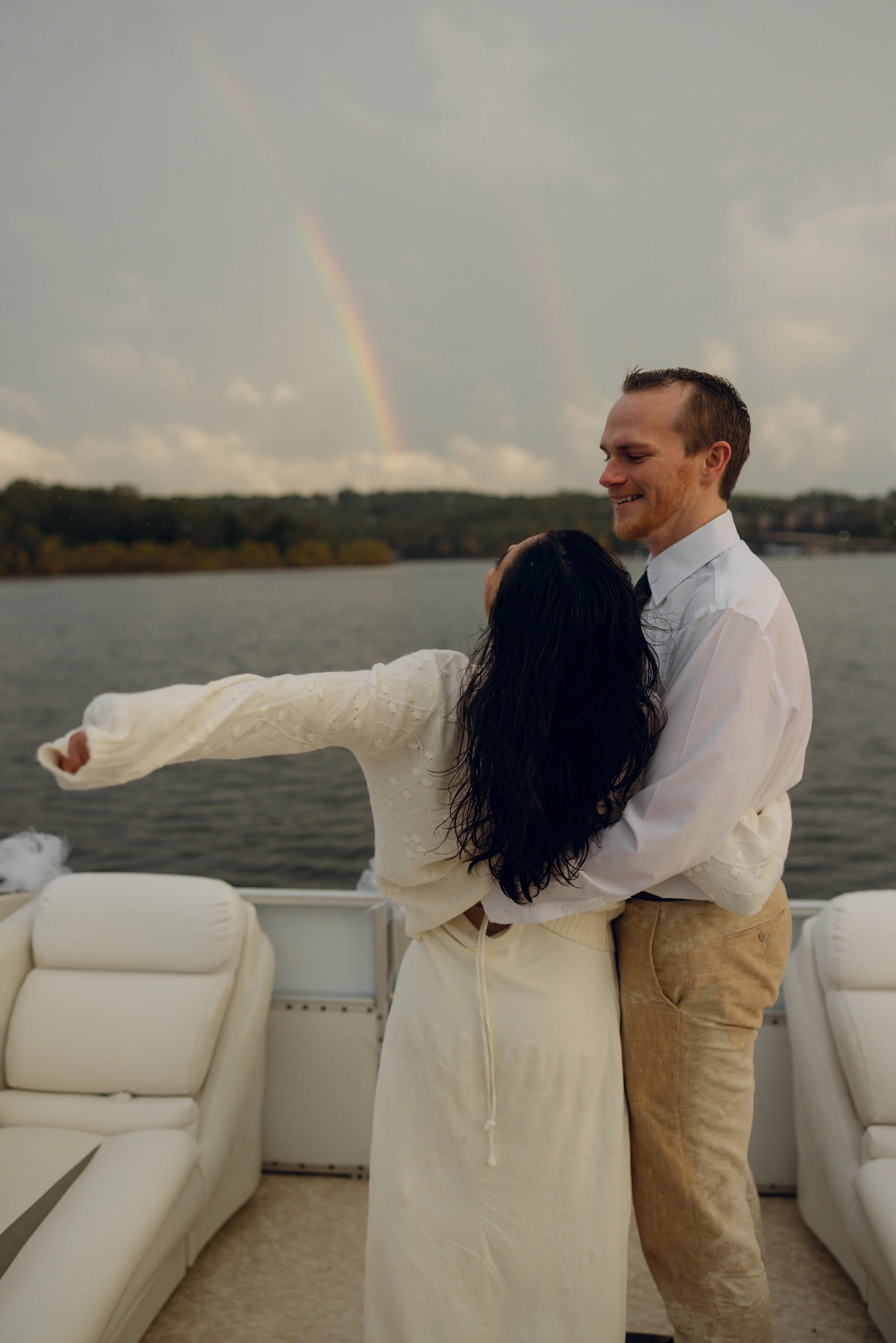 A couple dancing on a boat near a body of water with a rainbow in the cloudy sky.