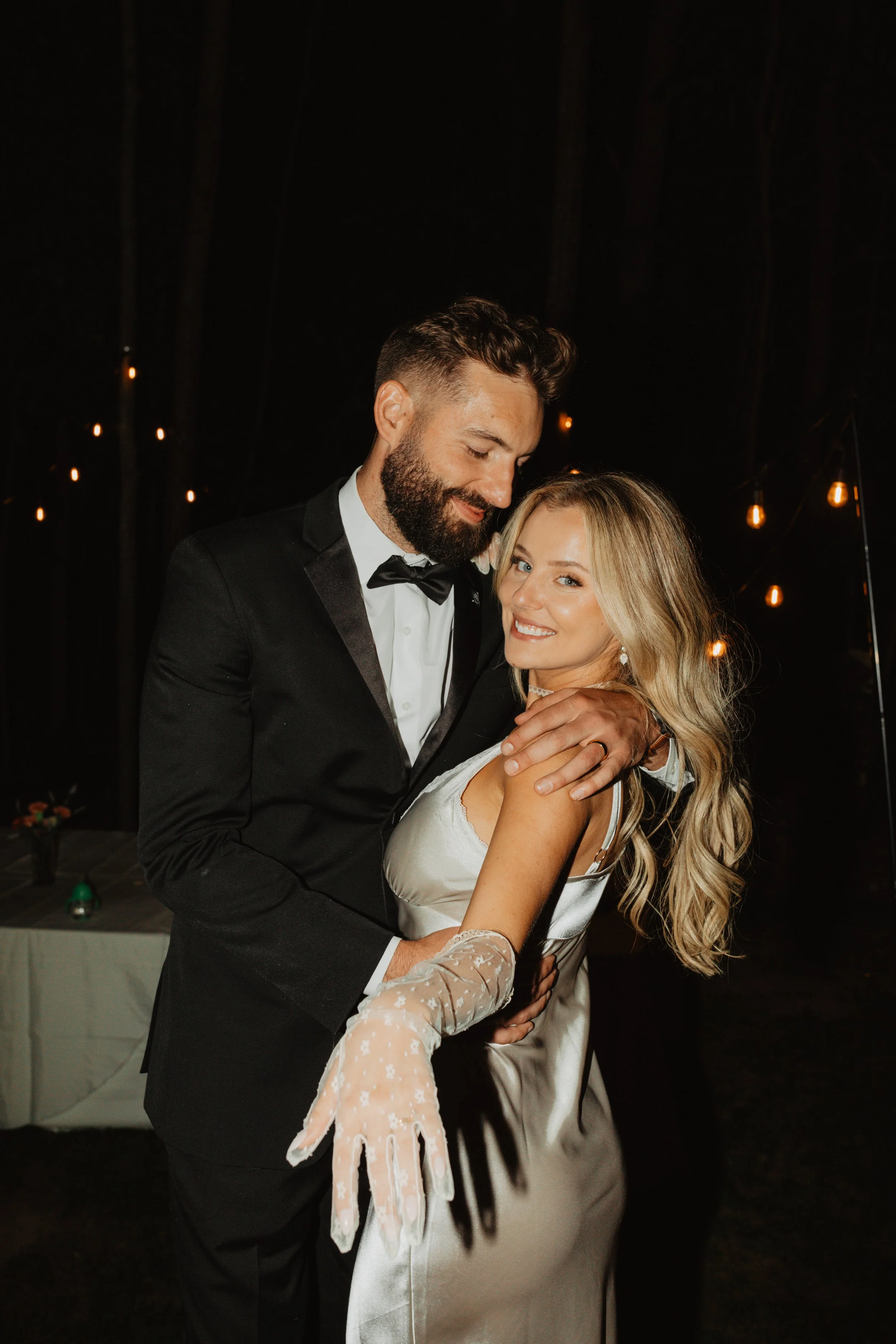 A man and a woman dancing at a wedding reception, with string lights in the background. The man is wearing a tuxedo, and the woman is in a satin wedding dress with lace gloves.
