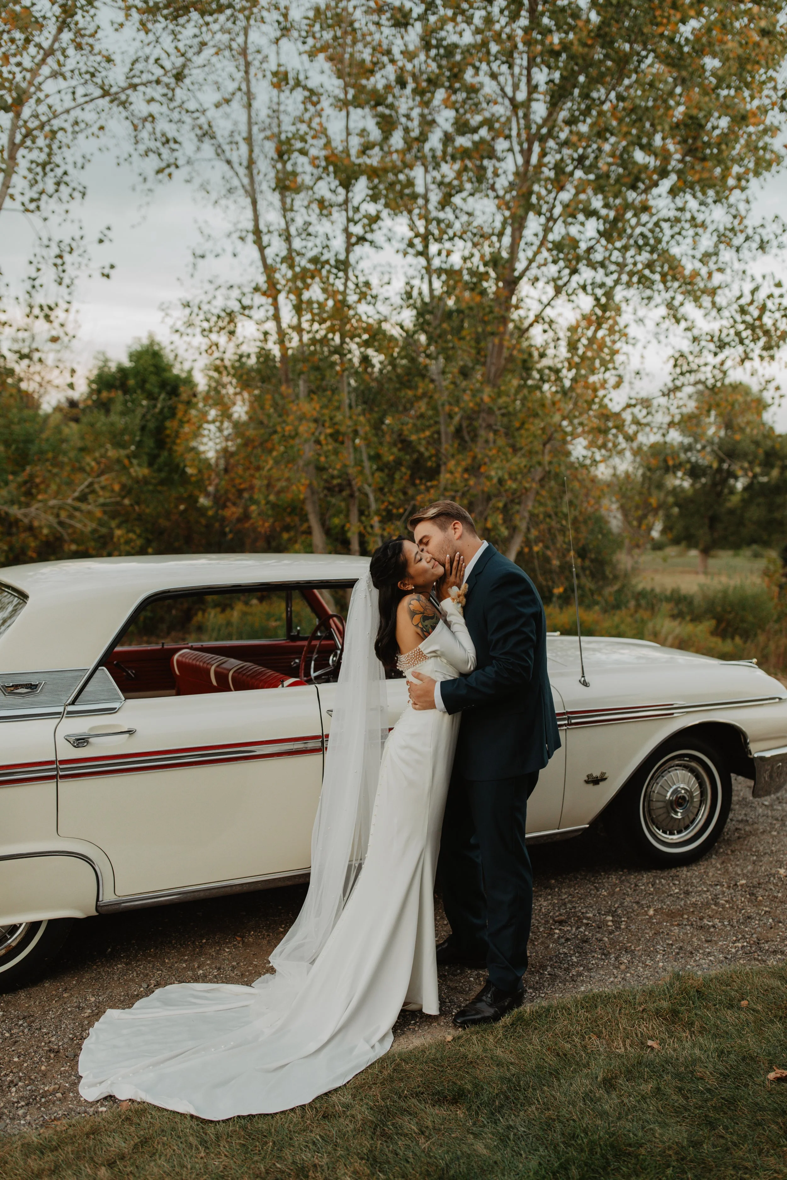 A bride and groom sharing a kiss in front of a vintage white car, surrounded by trees with autumn leaves.