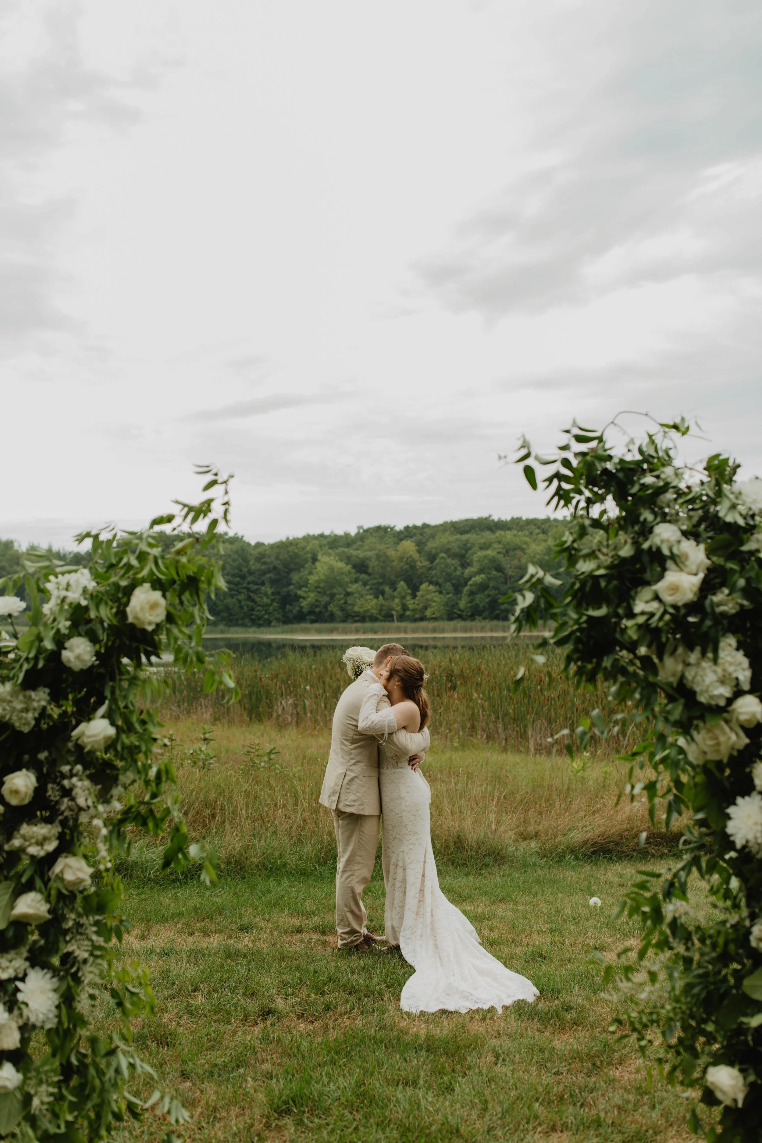 A bride and groom share a kiss during their outdoor wedding, framed by green floral arrangements, with a lake and forest in the background.