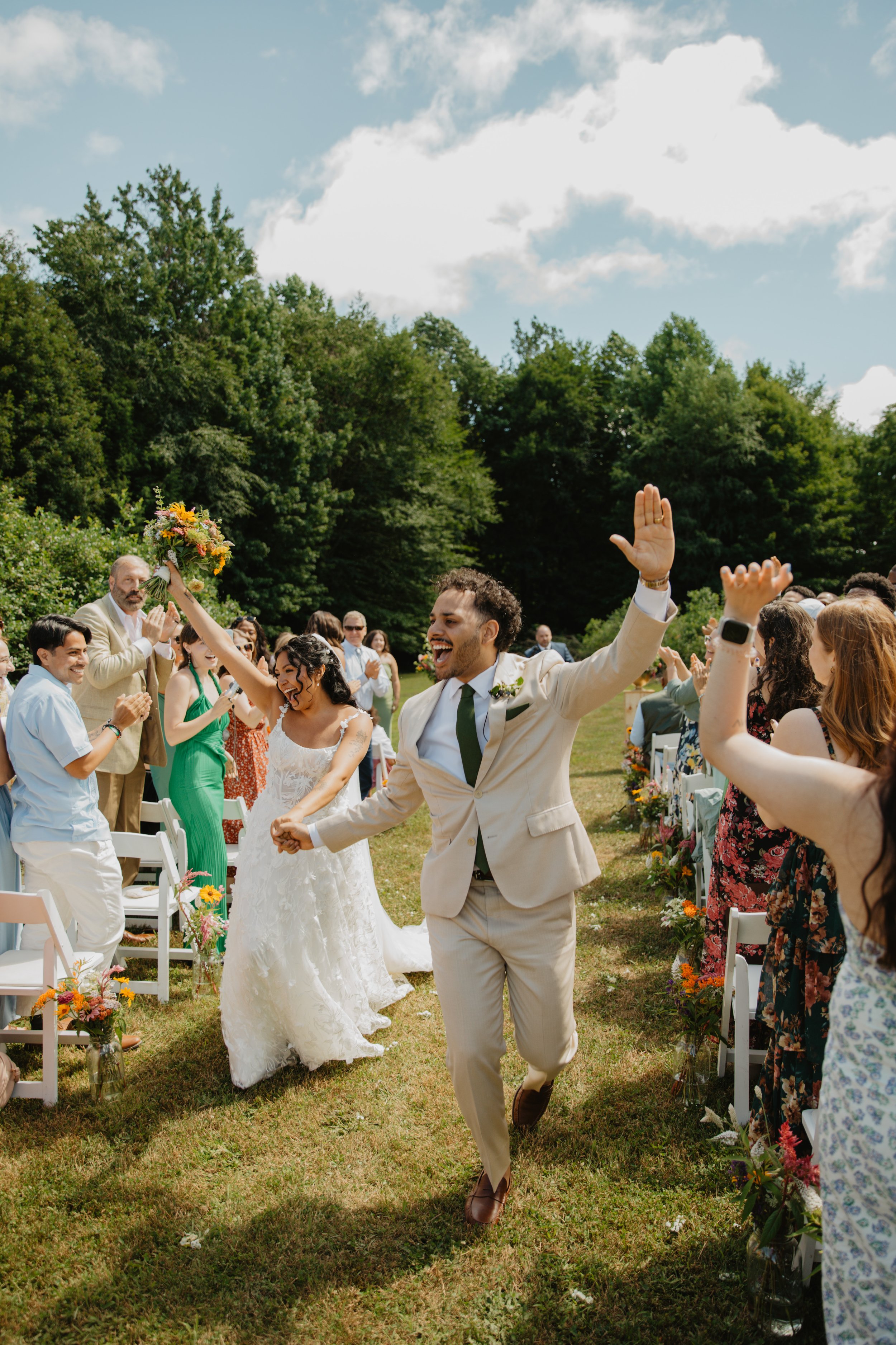 A joyful groom running down the aisle at an outdoor wedding, celebrating with guests clapping and cheering, in a green lawn setting with trees and a partly cloudy sky.