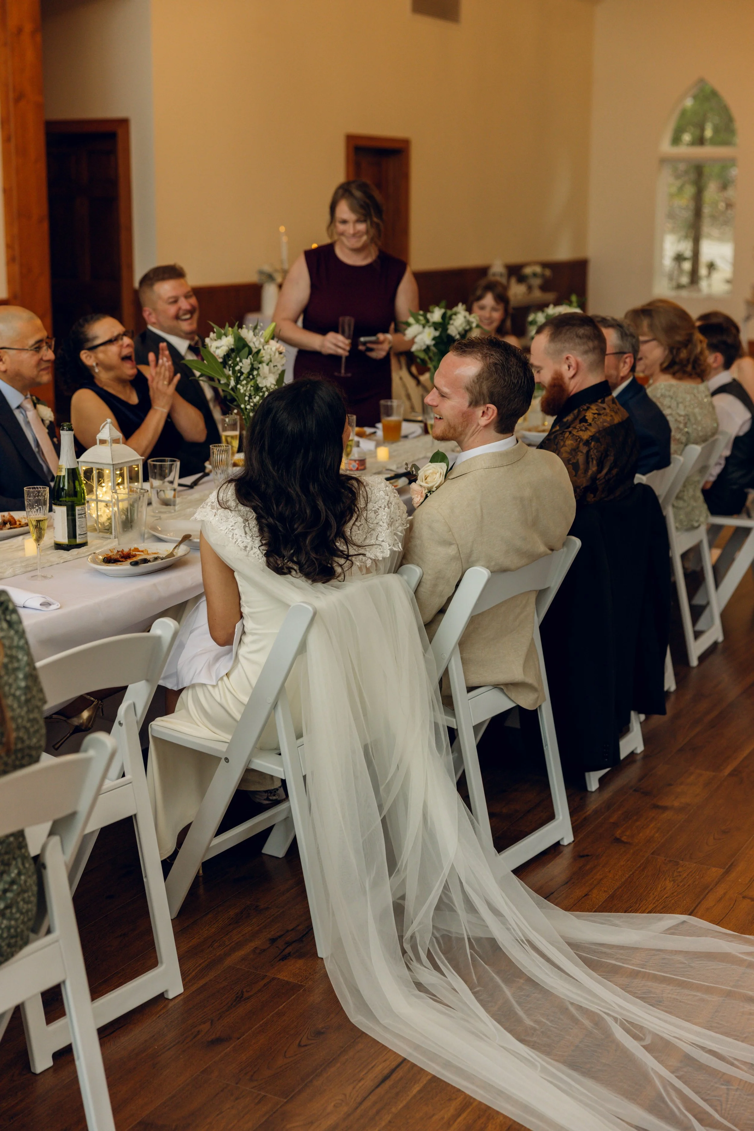 Bride and groom sitting at a wedding reception table, smiling at each other, surrounded by guests.
