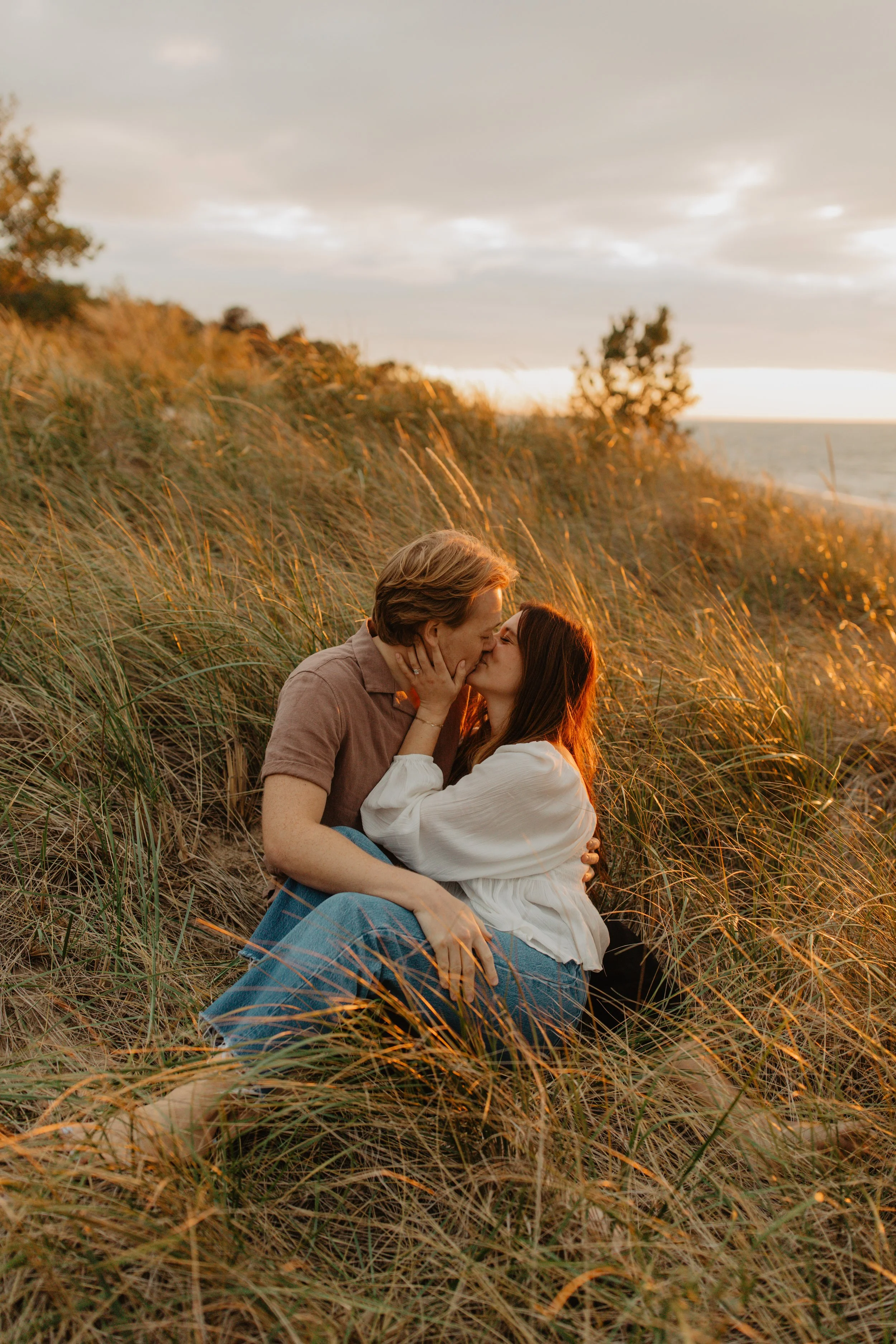 A couple sharing a kiss on a beach at sunset surrounded by tall grass.