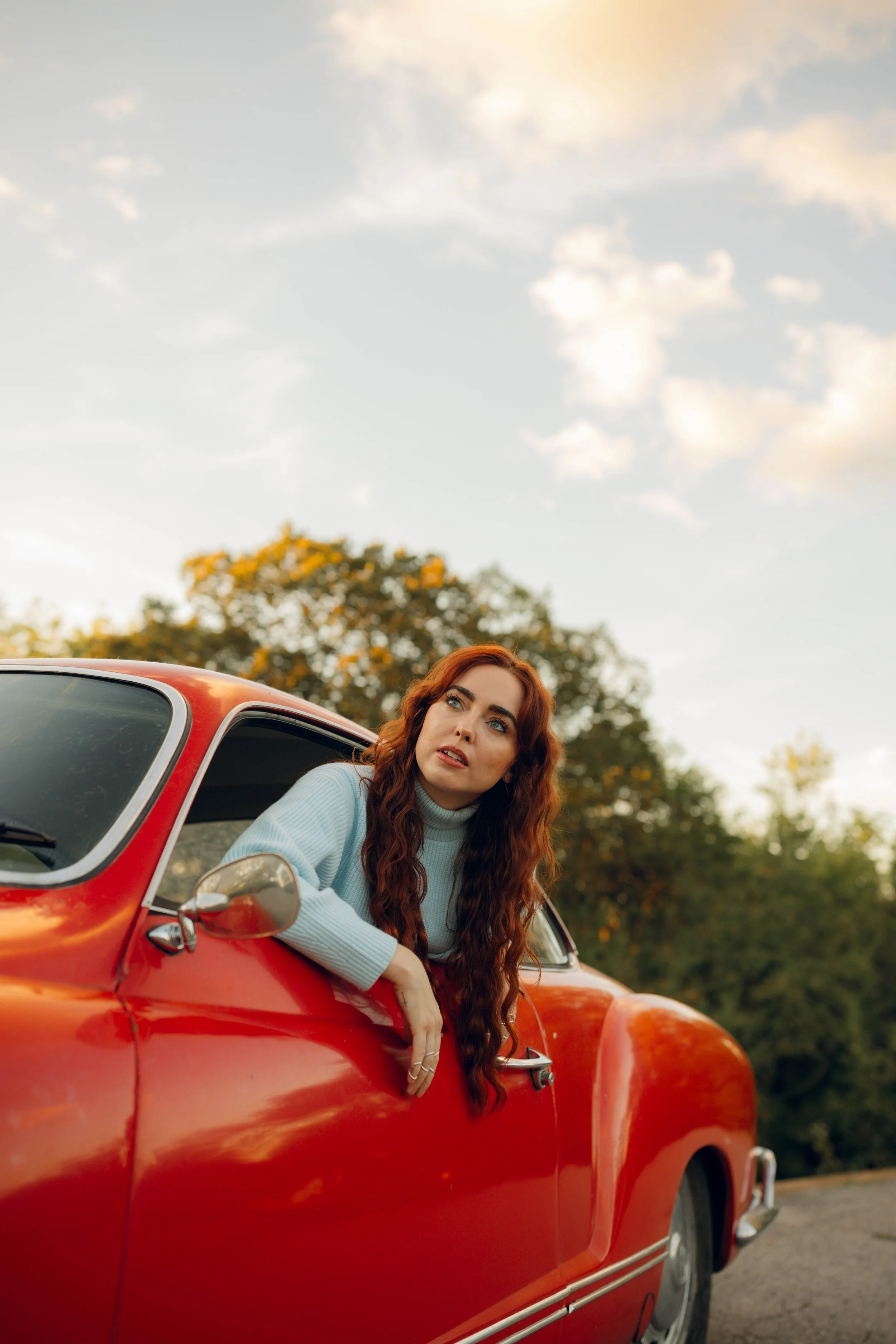 A woman with long, wavy red hair leaning out of the window of a red vintage car, looking into the distance with a thoughtful expression, outdoors during sunset.