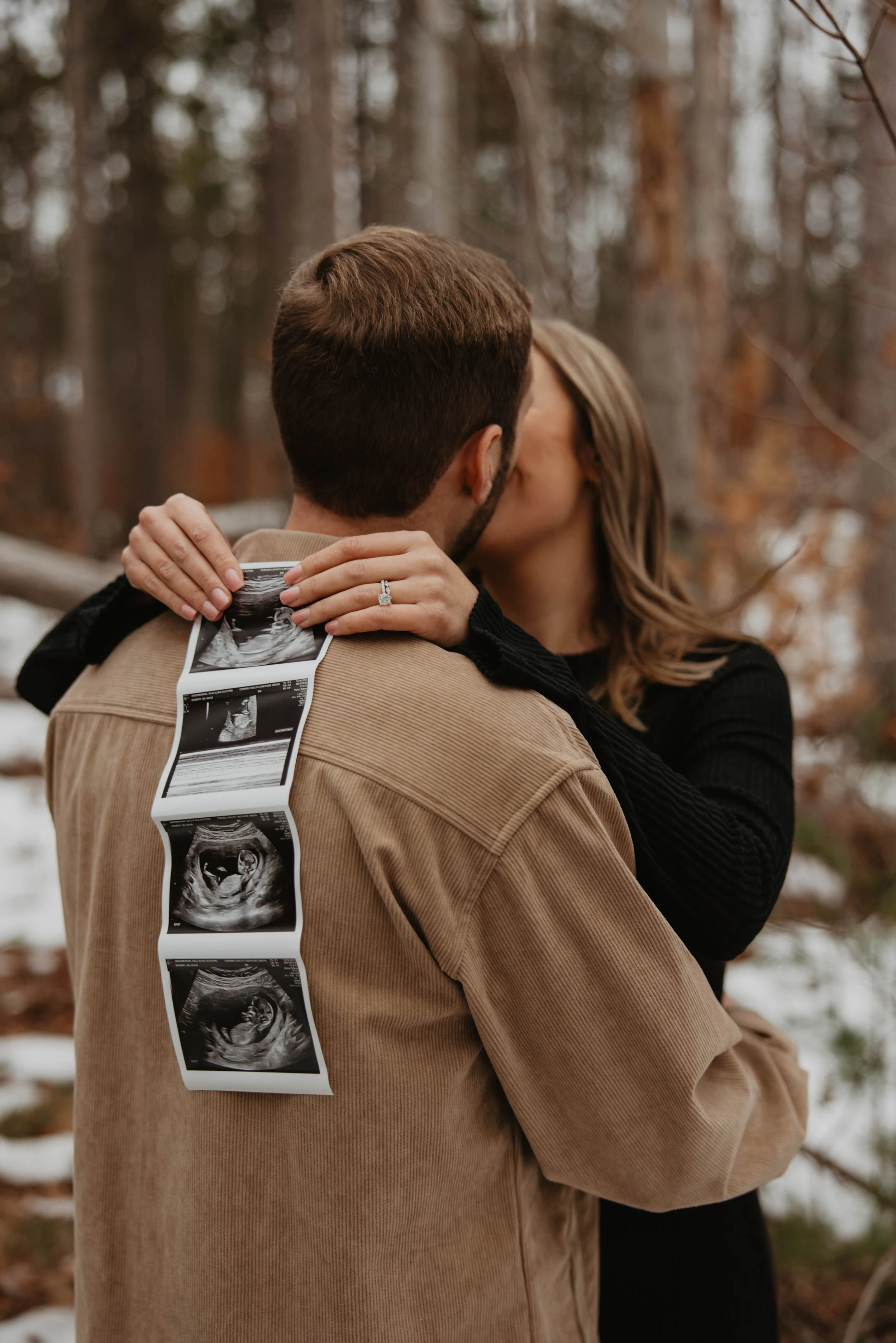 A couple kissing outdoors in a wooded area with snow on the ground. The woman is holding an ultrasound photo on the man's shoulder, revealing a fetus.