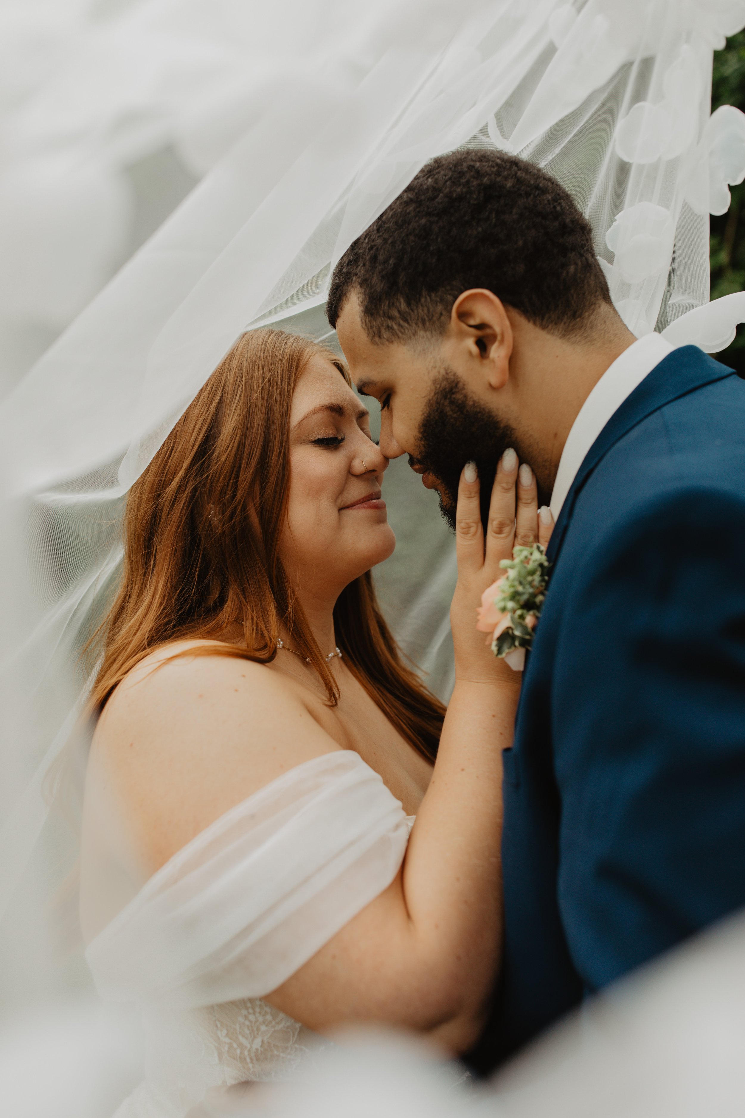 A bride and groom with their foreheads touching, holding each other's faces, smiling, with a sheer white veil or fabric draped around them.