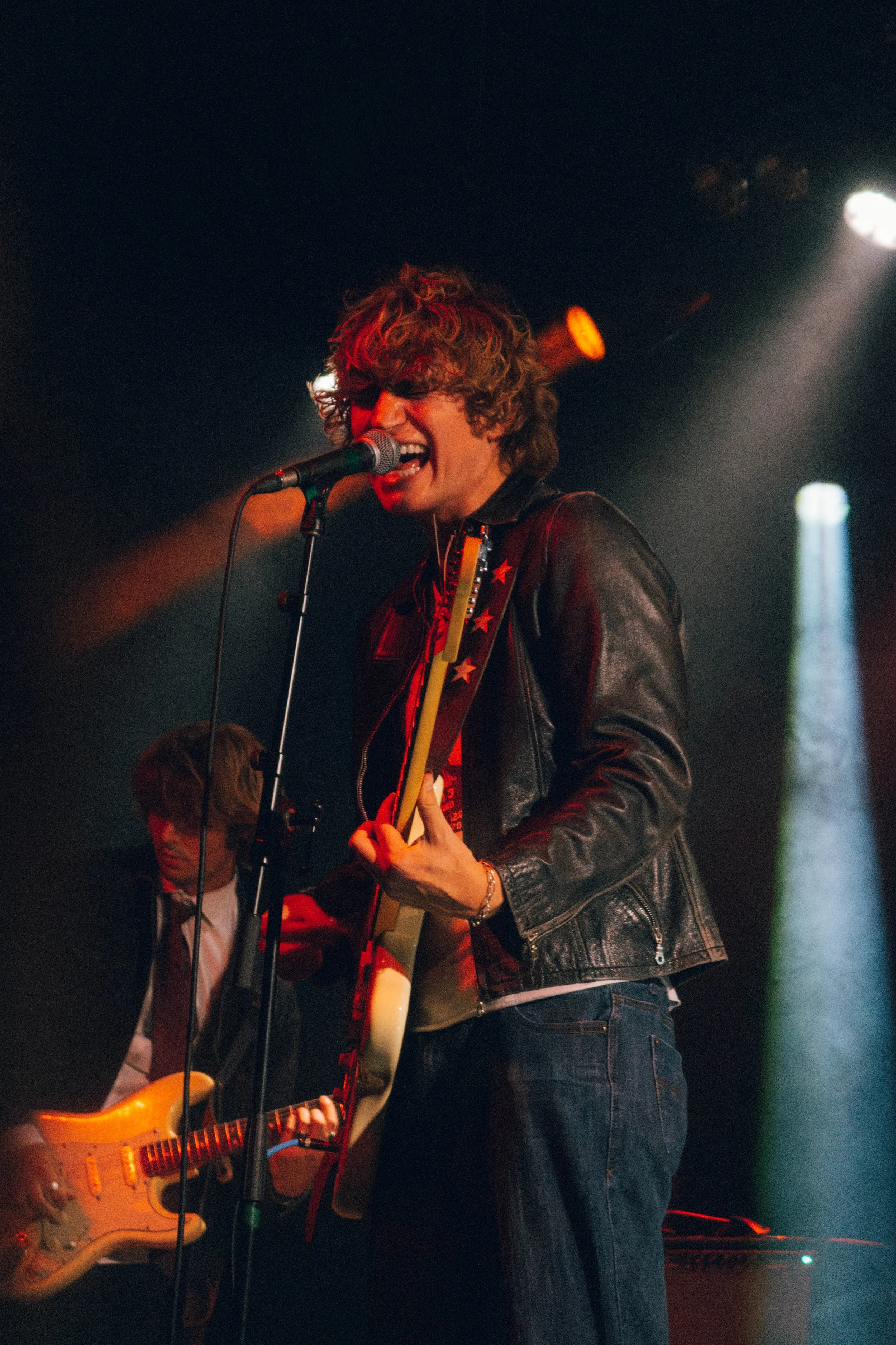 The band SUNROOM singing into a microphone while playing an electric guitar on stage, wearing a black leather jacket at a concert in Chicago.