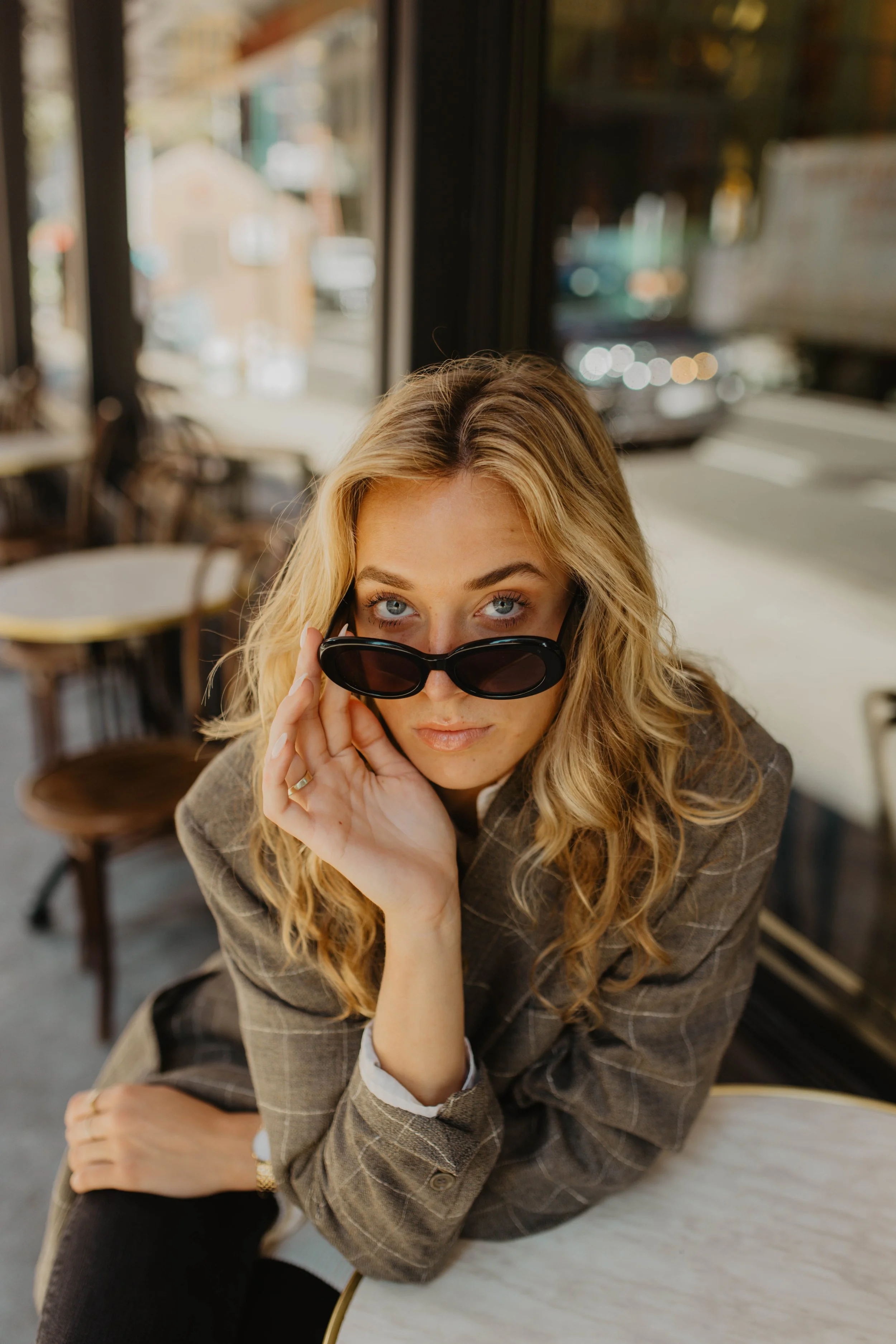 A woman with blonde curly hair wearing a brown plaid blazer and black sunglasses, sitting at a table in a cafe, looking over her sunglasses at the camera.