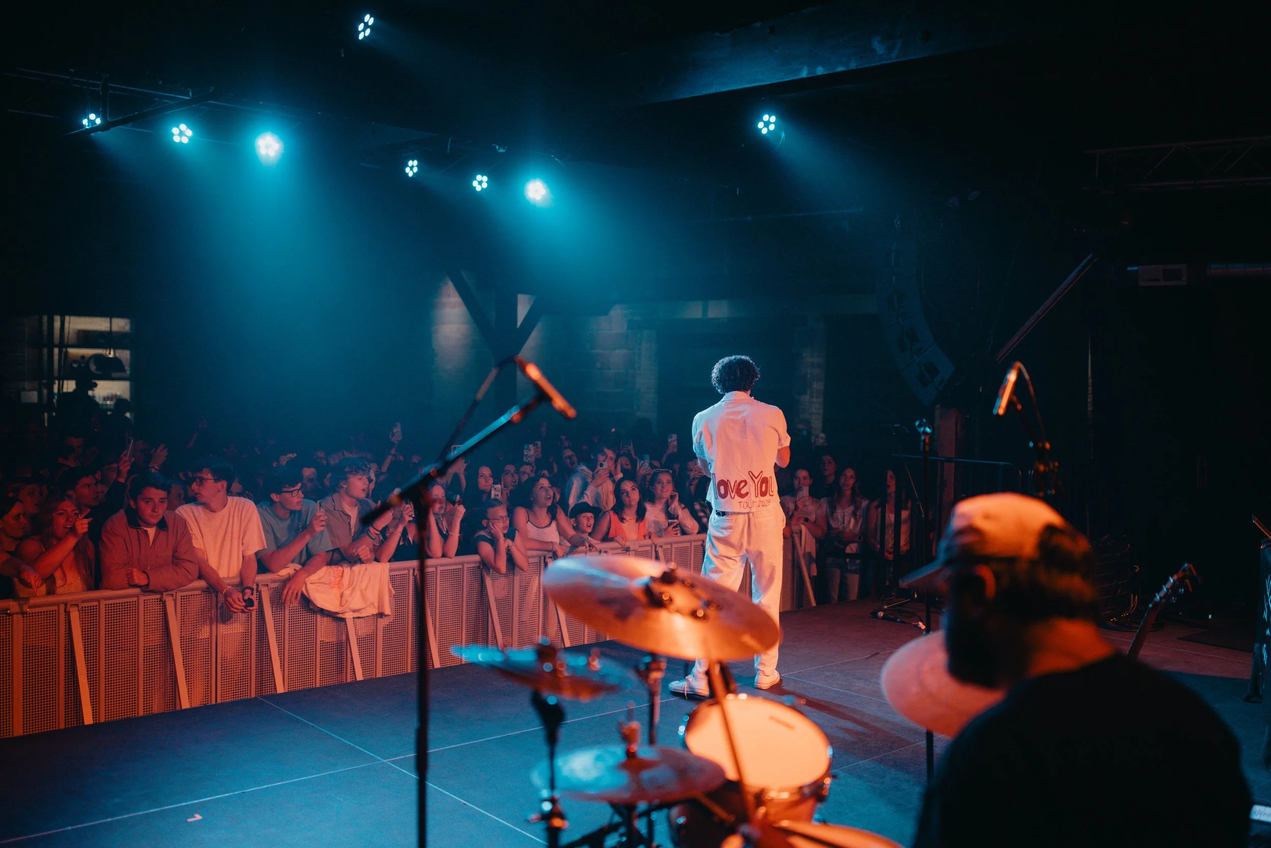 Sammy Rash on stage facing an audience at a concert, with a drummer in the foreground, and stage lights overhead.