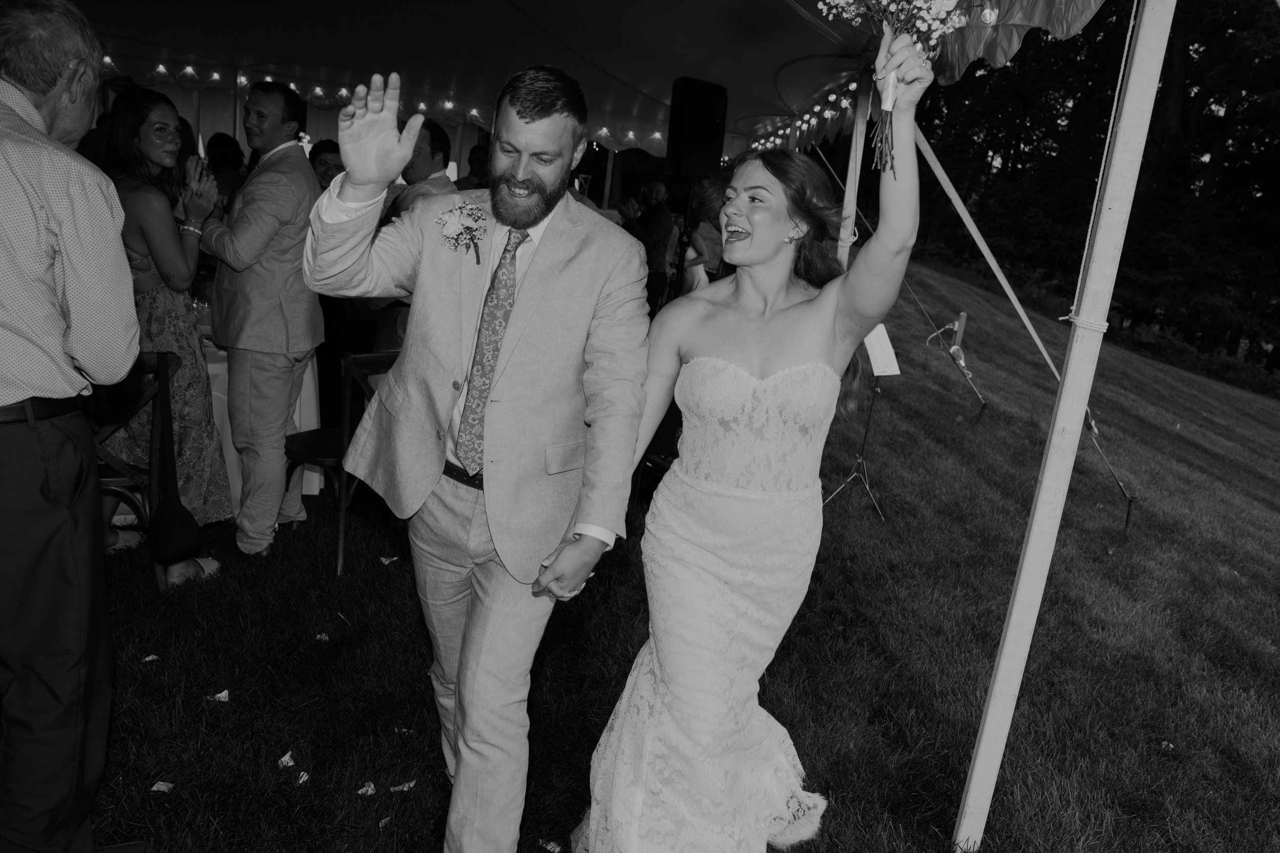 Wedding couple dancing at outdoor reception, woman in white lace gown holding bouquet, man in light suit with floral boutonniere, guests around under decorated tent.