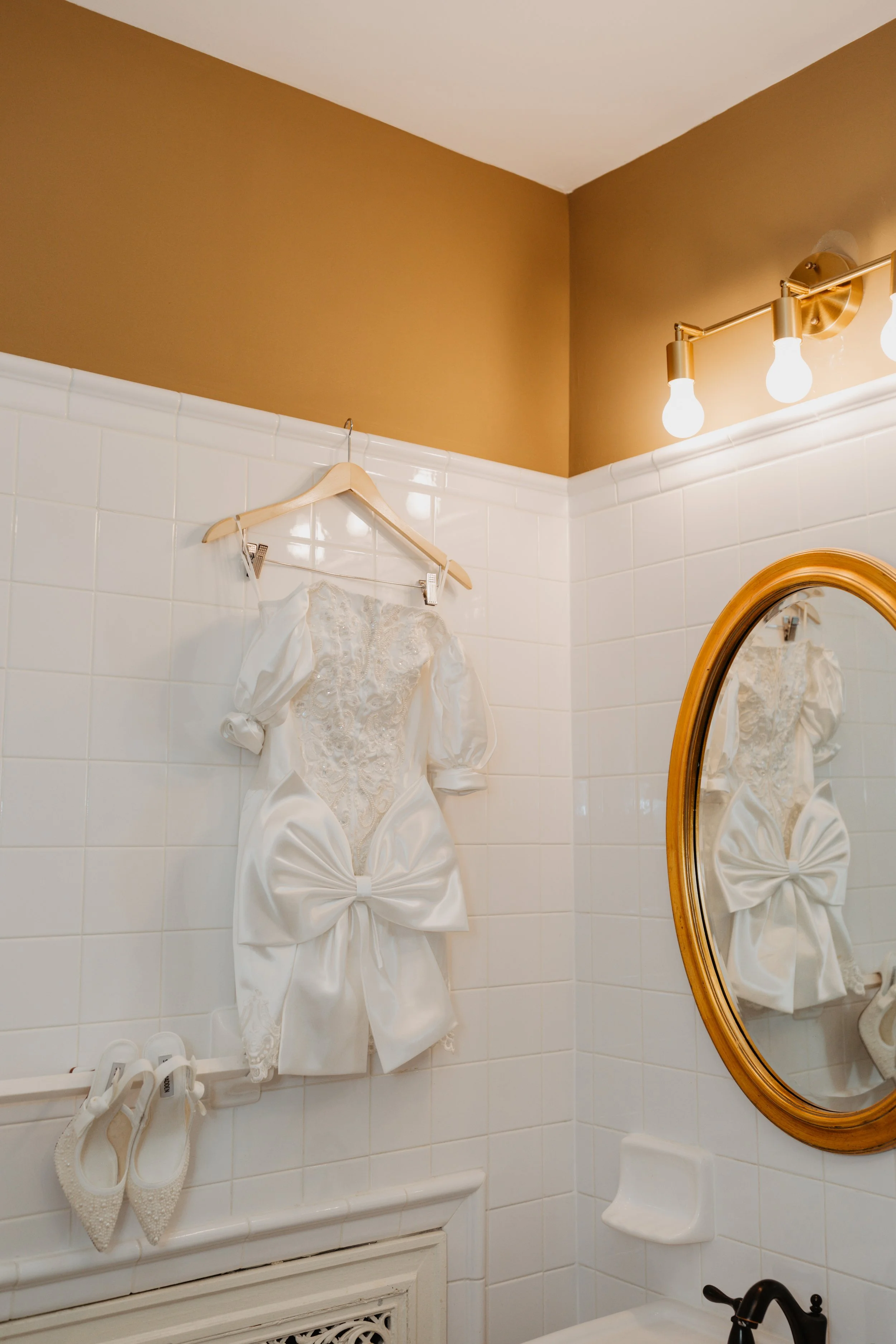 A white wedding dress with puffed sleeves and a large bow at the waist is hanging on a hanger inside a bathroom with white tiled walls and brown upper walls. A pair of white shoes is placed below the dress. There is a mirror on the wall reflecting th